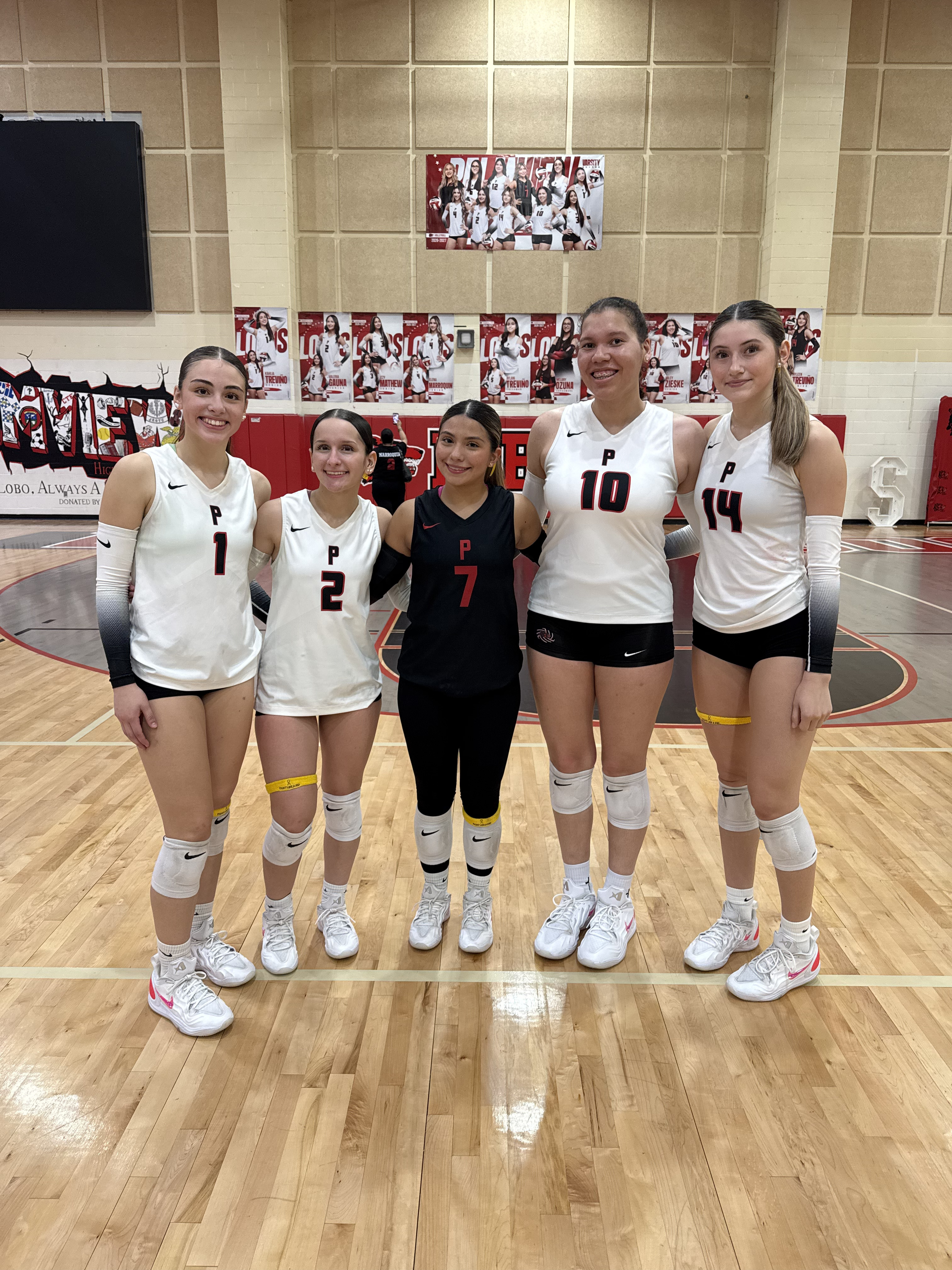 Five female volleyball players in uniforms stand smiling on a polished wooden court in a gymnasium.
