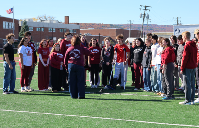 A diverse choir of high school students sings on an outdoor athletic field, directed by a conductor in a maroon jacket.