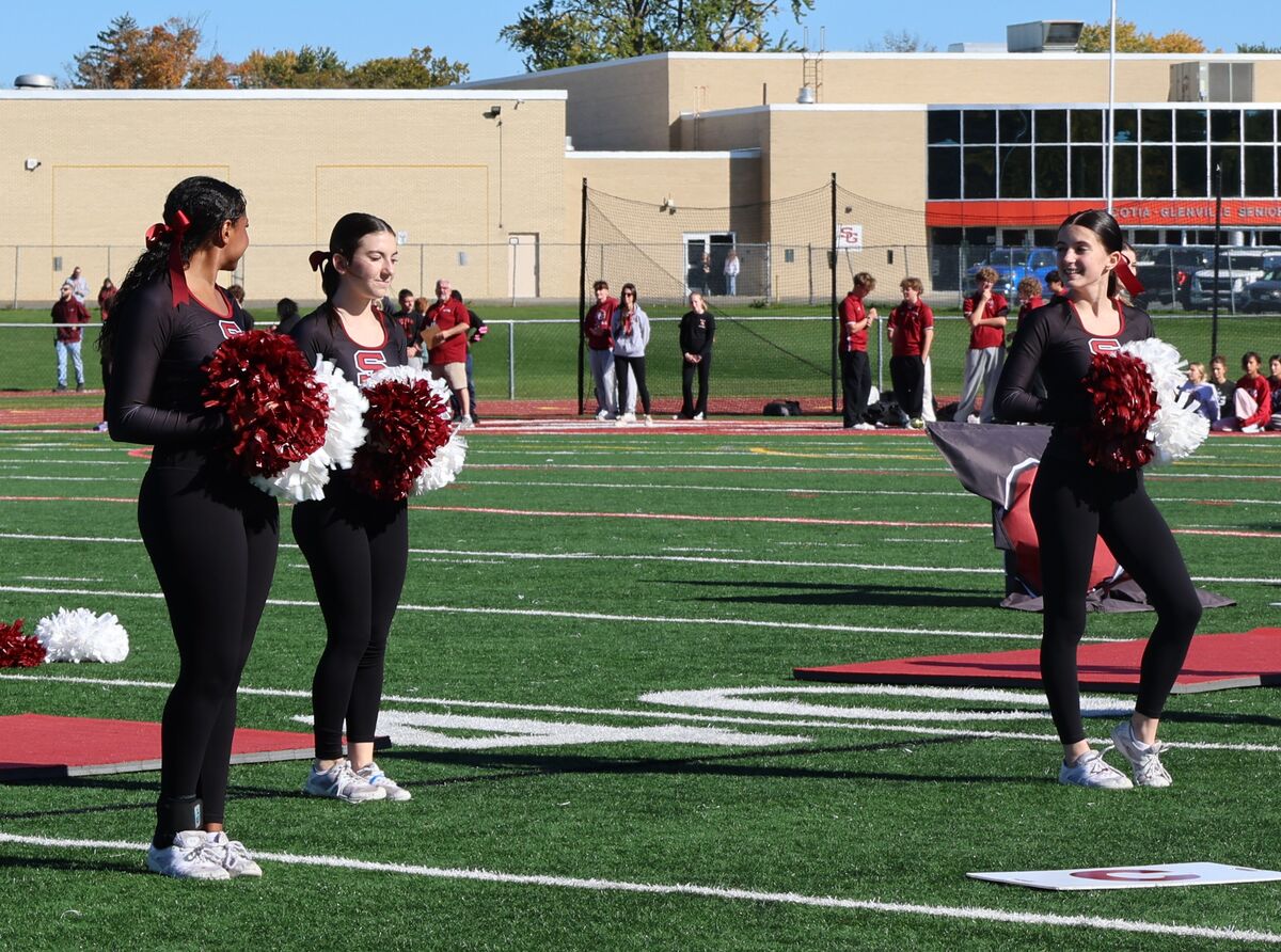 Three cheerleaders in black uniforms and red and white pom-poms stand on a sunny athletic field.