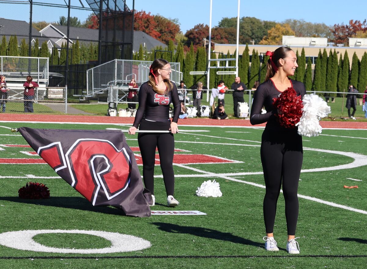 Two high school cheerleaders in black uniforms stand on a green athletic field.