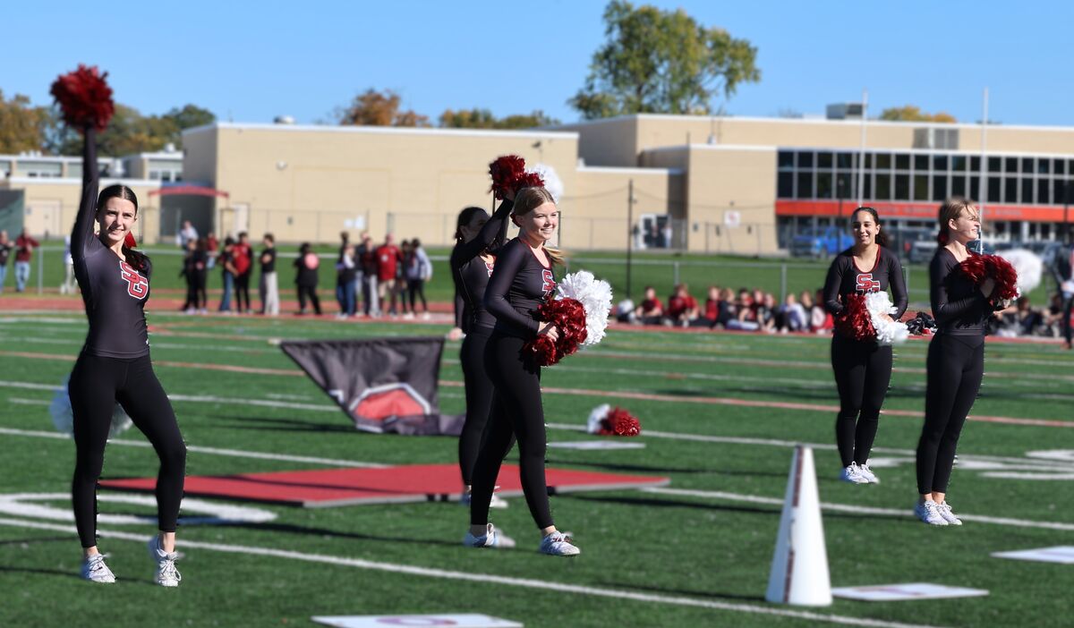 A group of cheerleaders in black uniforms and maroon and white pom-poms perform on a green football field.