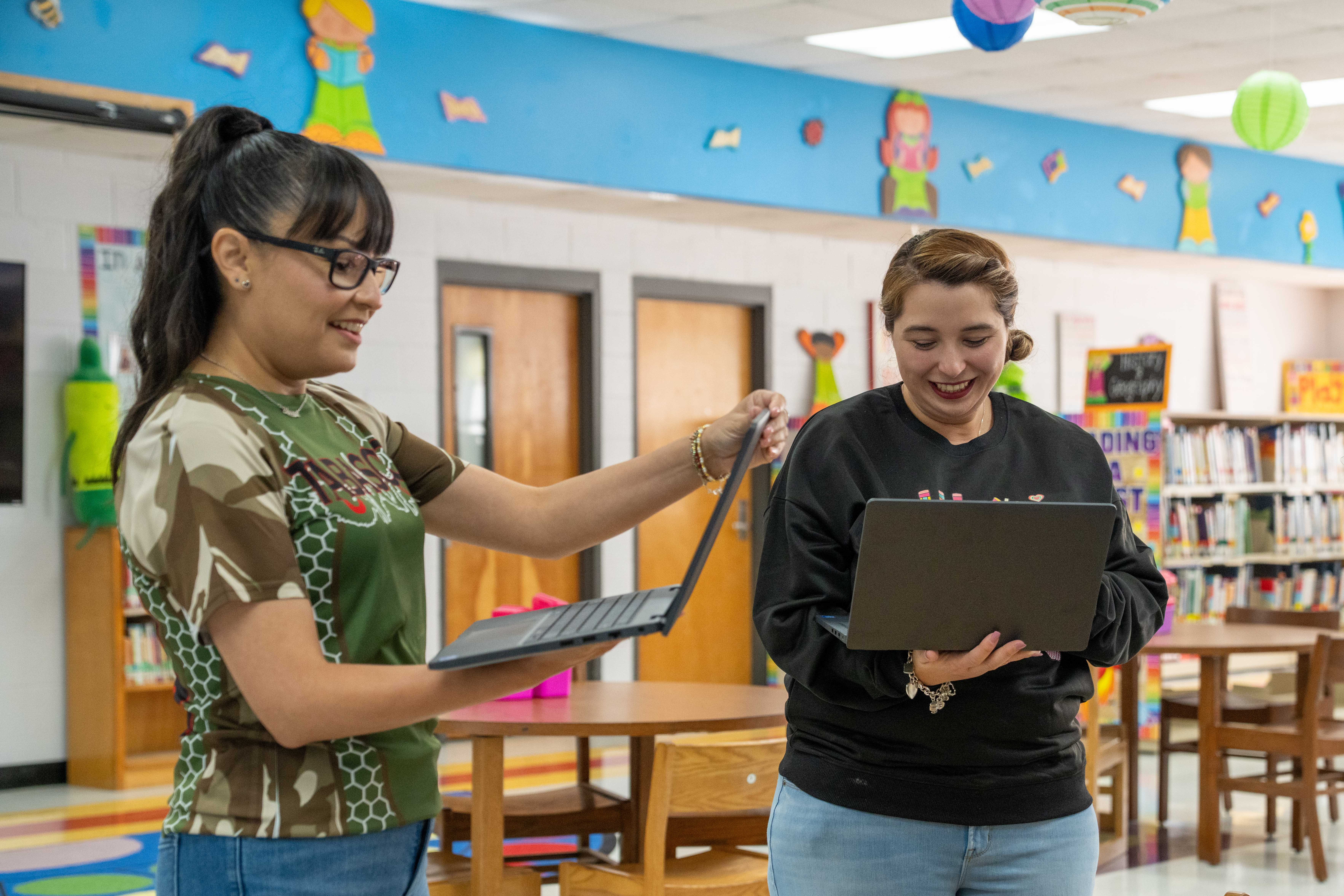 Two women stand in a colorful school library, smiling as they look at their laptops.