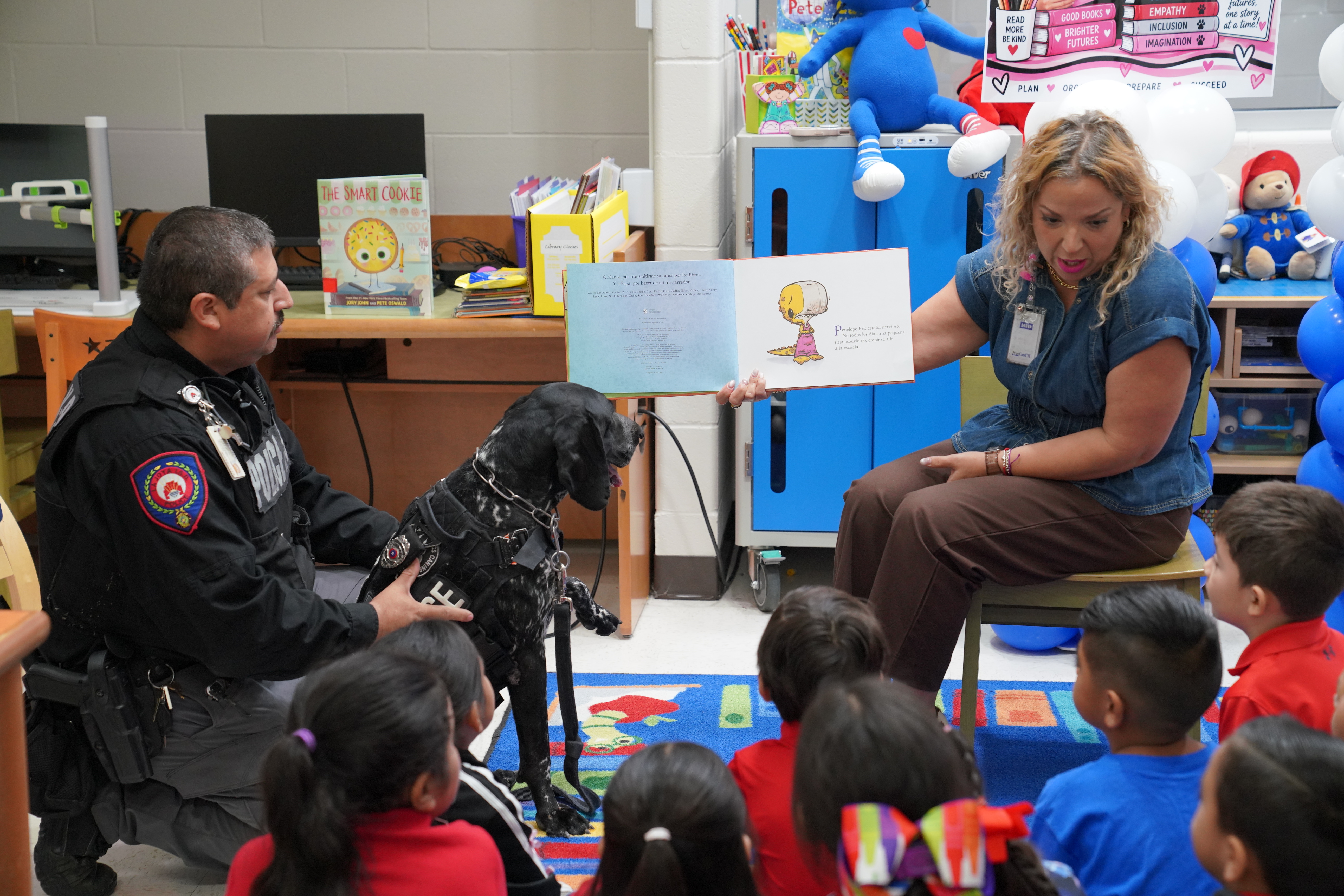 A woman reads a book to a group of children in a classroom while a police officer and a dog sit nearby.