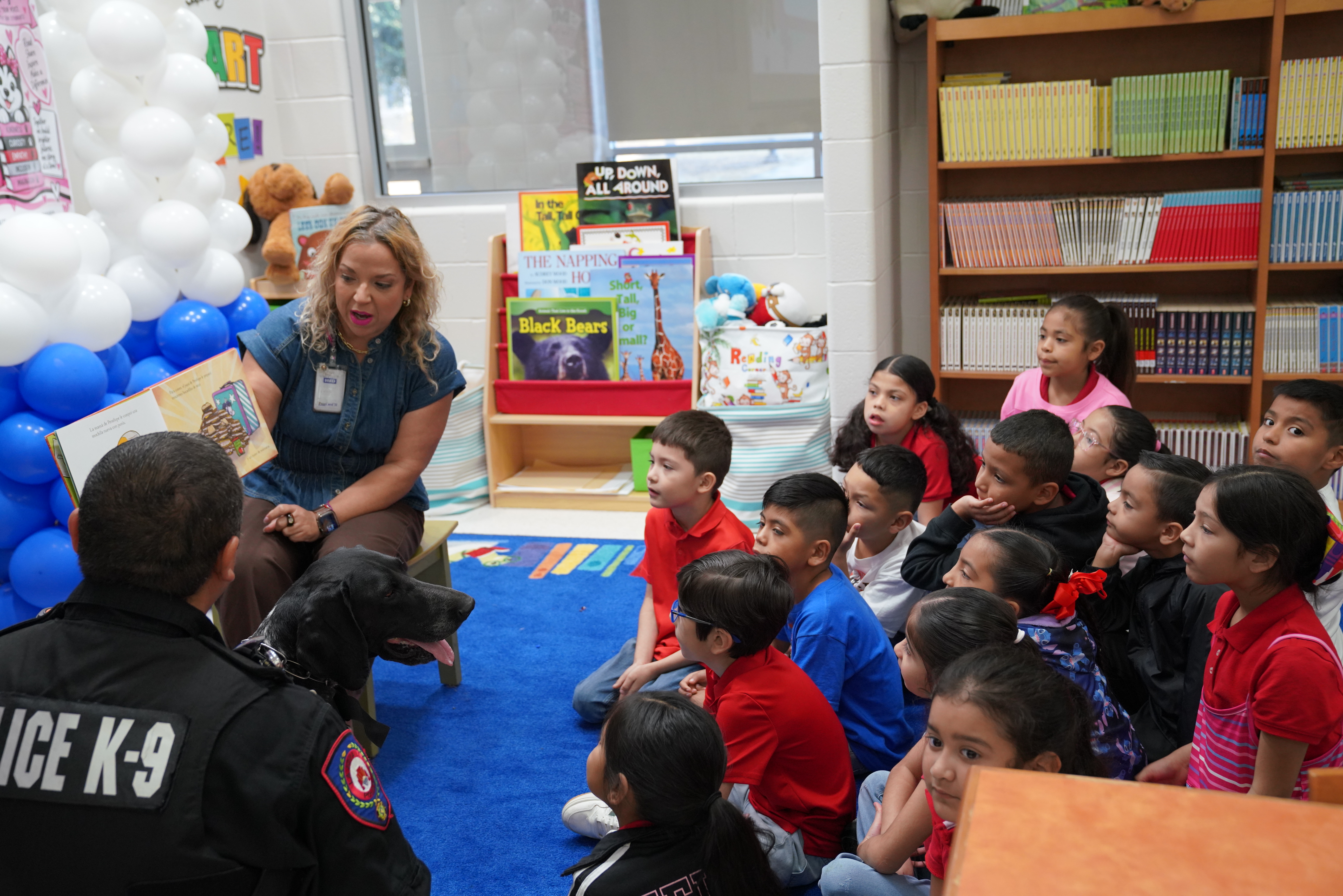 A teacher reads a story to a group of attentive children sitting on a blue rug, with a police officer and a K-9 dog present.