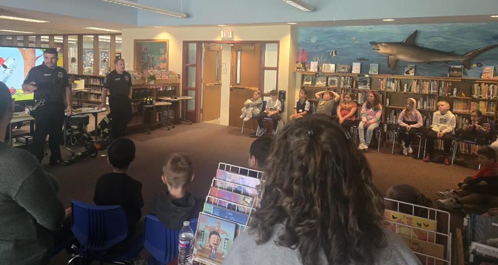 Two firefighters stand before a group of seated children in a library, presenting a community safety talk.