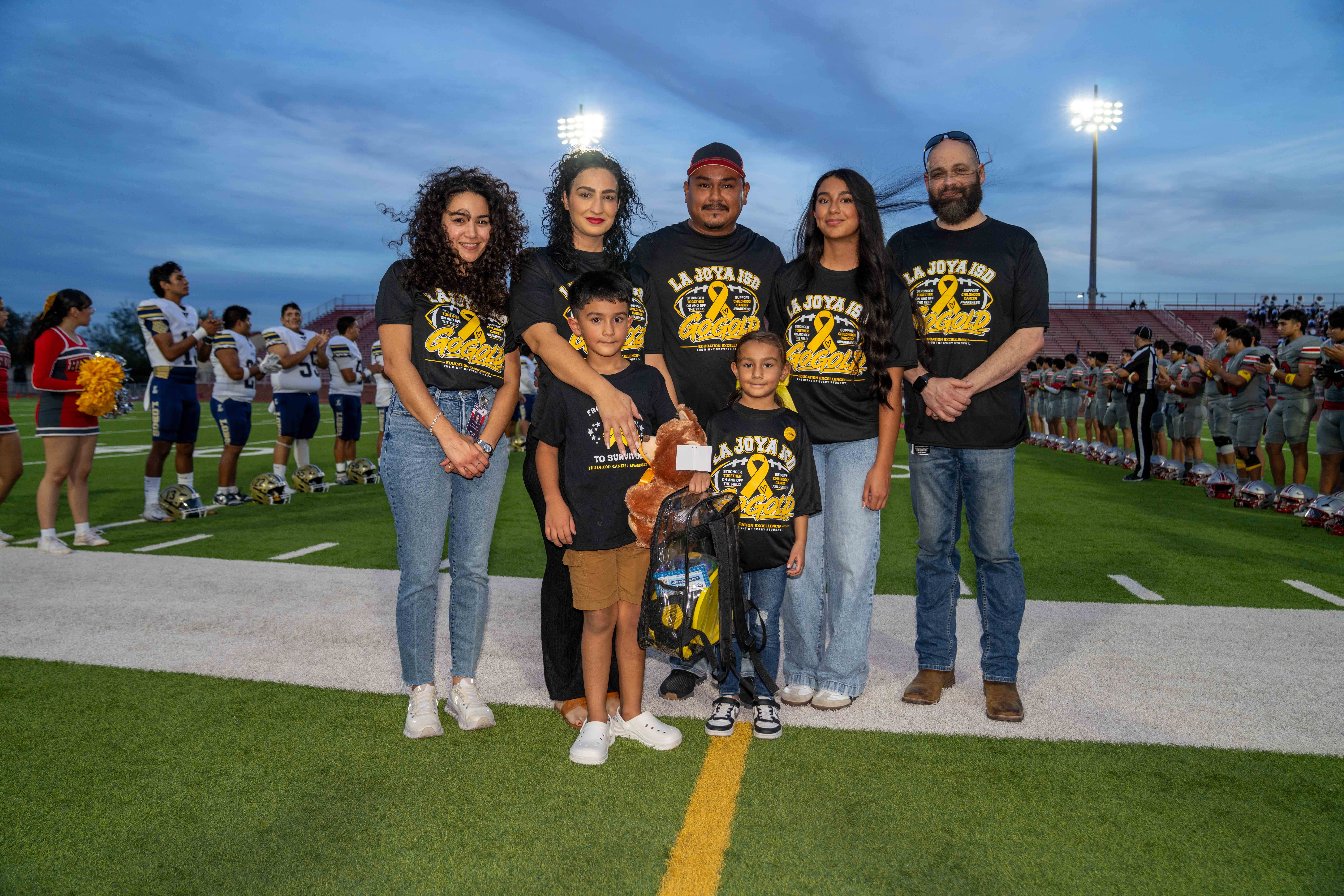 A group of adults and children stand together on a football field, wearing matching black t-shirts with yellow text and ribbons for childhood cancer awareness.