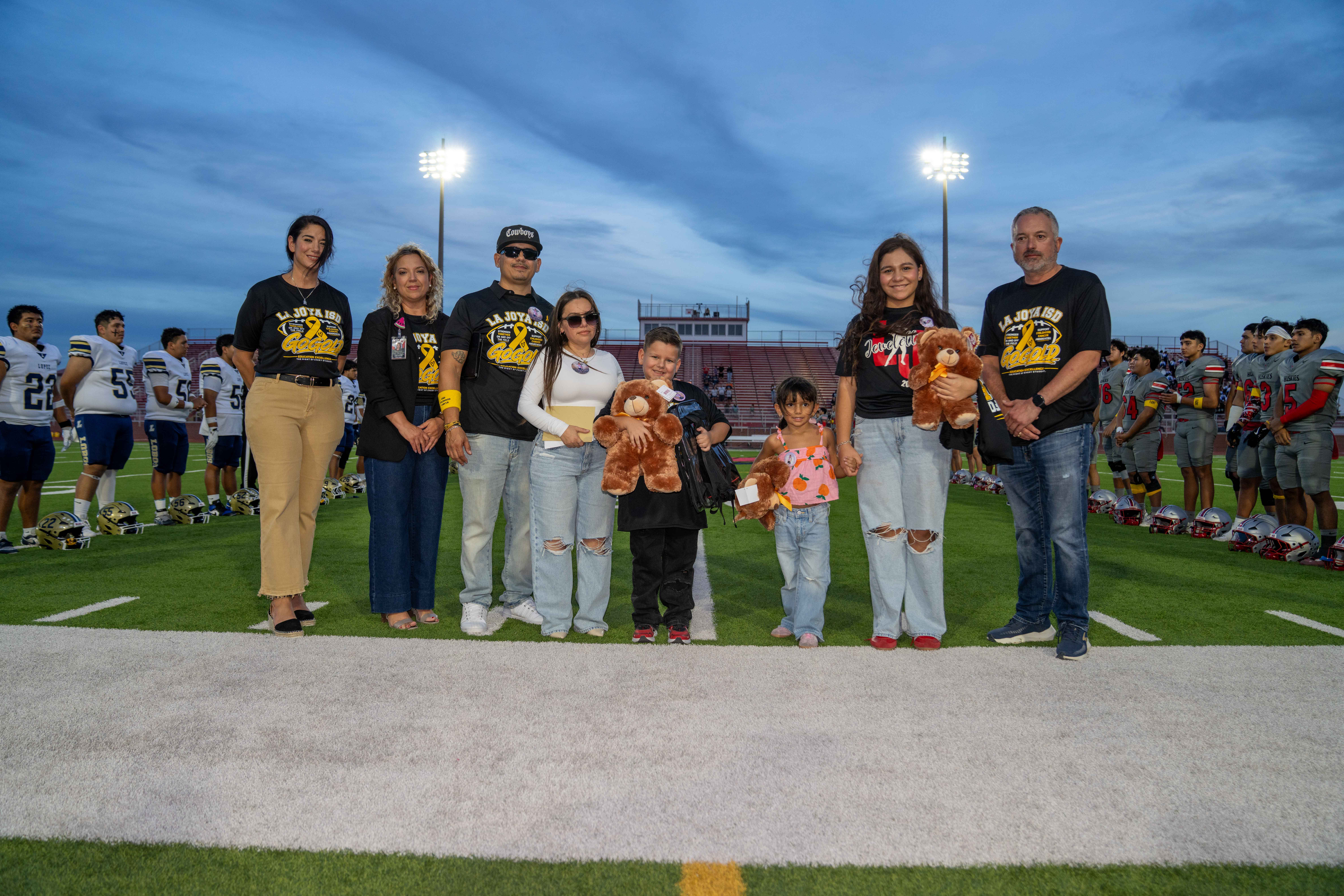 A group of adults and children stand proudly together on a football field holding teddy bears.