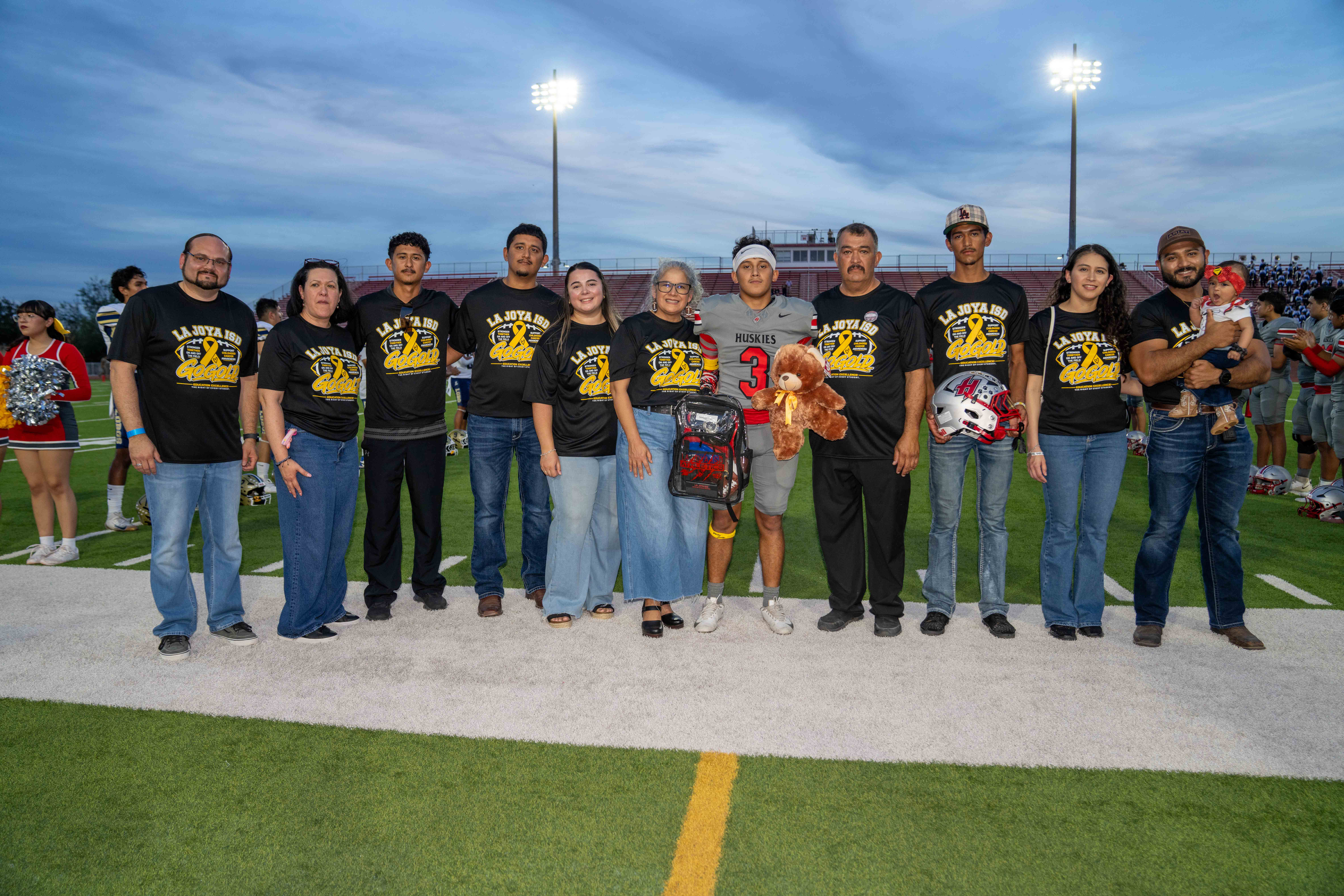 A diverse family and football player stand together on a lit-up turf field, smiling for a group photo wearing matching shirts.