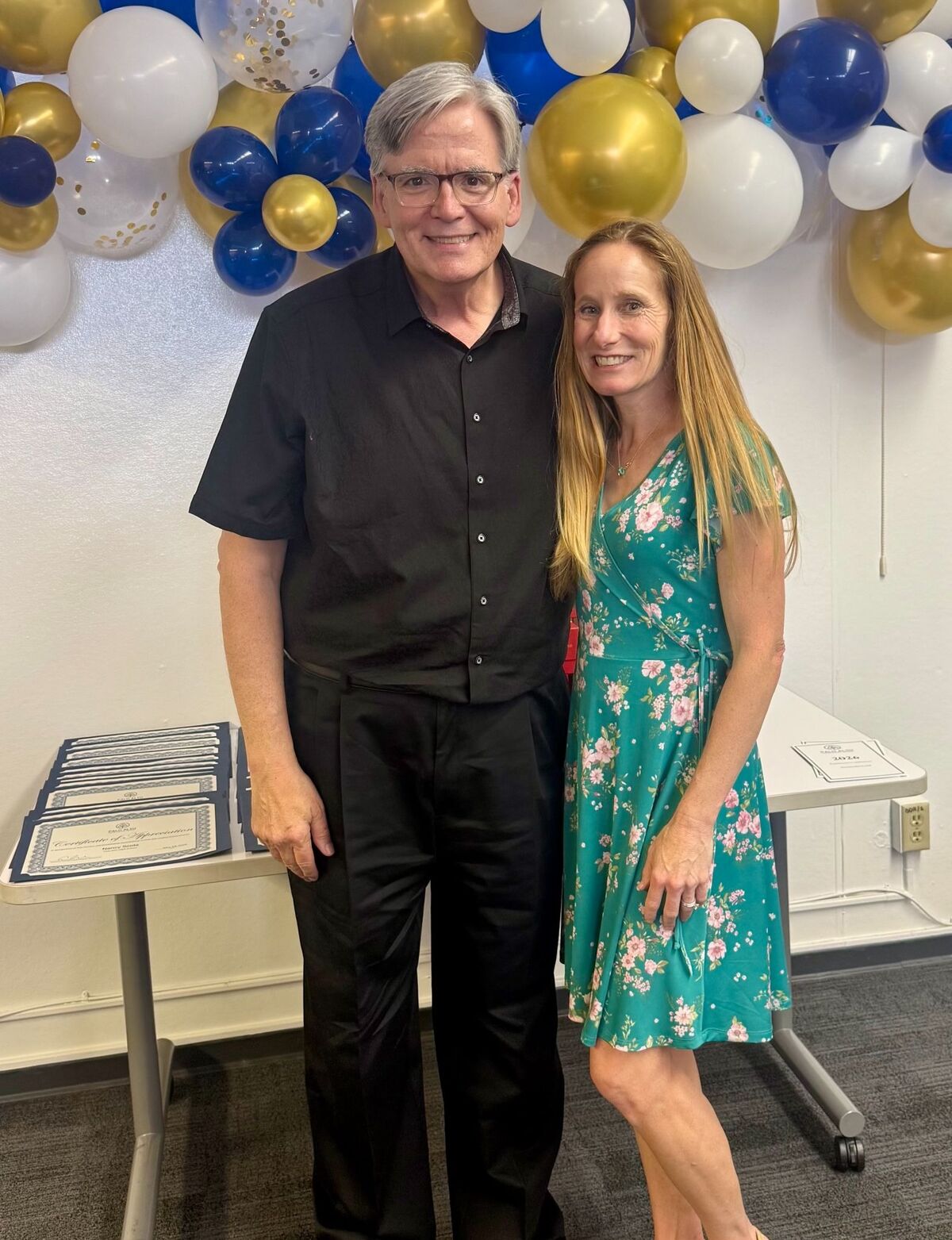 A man and a woman smile in front of a balloon arch, with certificates on a table.