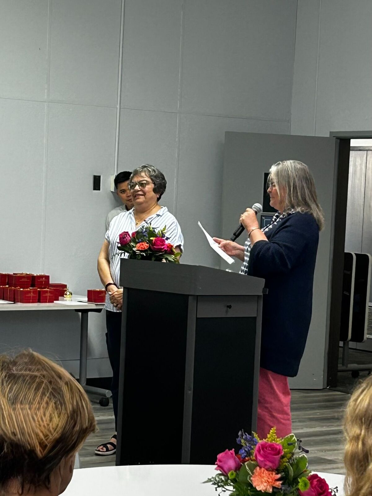 A woman speaks into a microphone at a podium, holding a paper, with a bouquet of flowers.