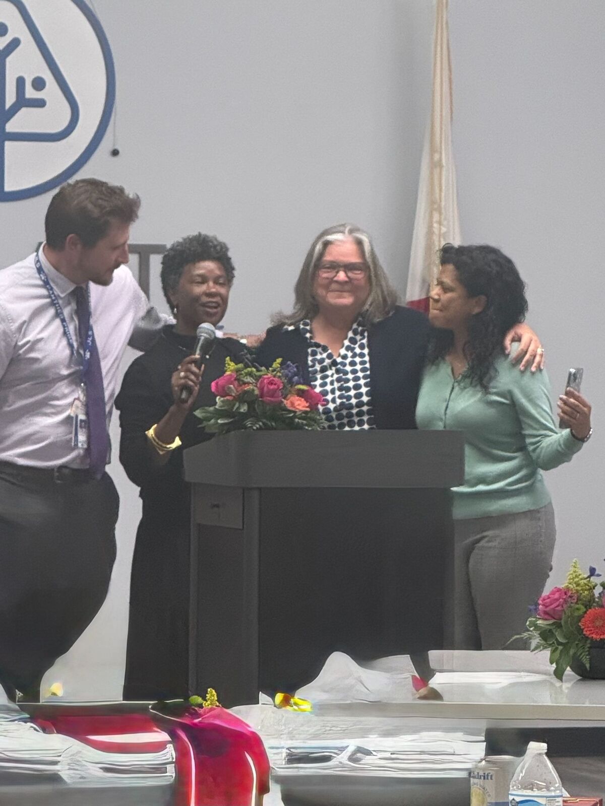 Four people stand around a podium with a microphone and a floral arrangement.