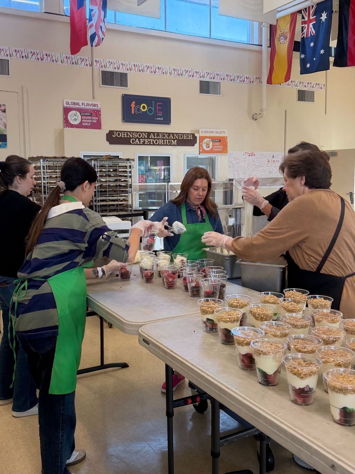 People in aprons prepare individual parfaits with fruit and granola in a cafeteria.