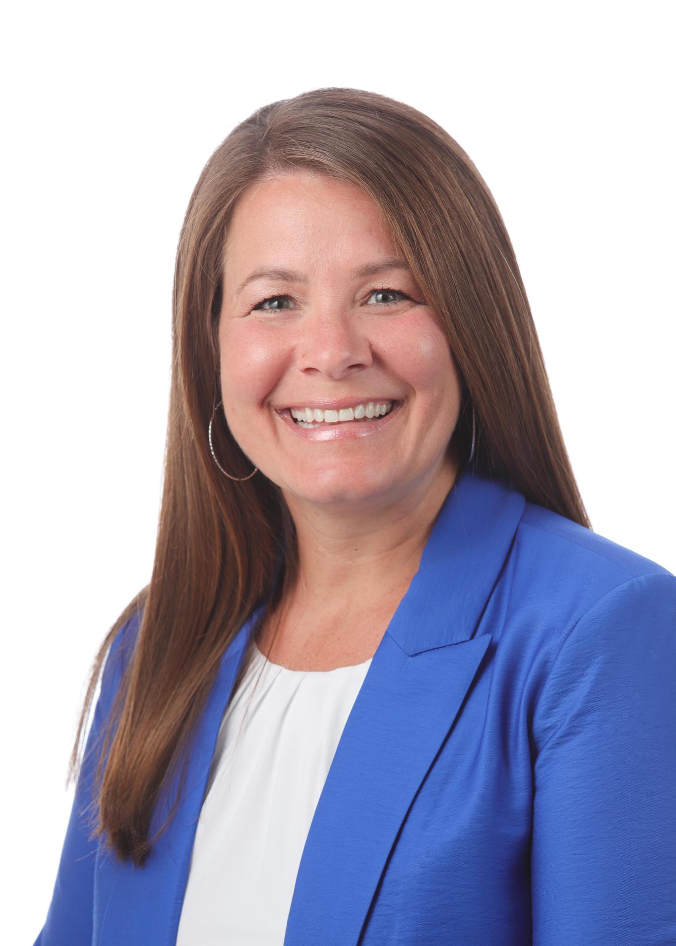 A woman with long brown hair smiles warmly, wearing a blue blazer over a white top.