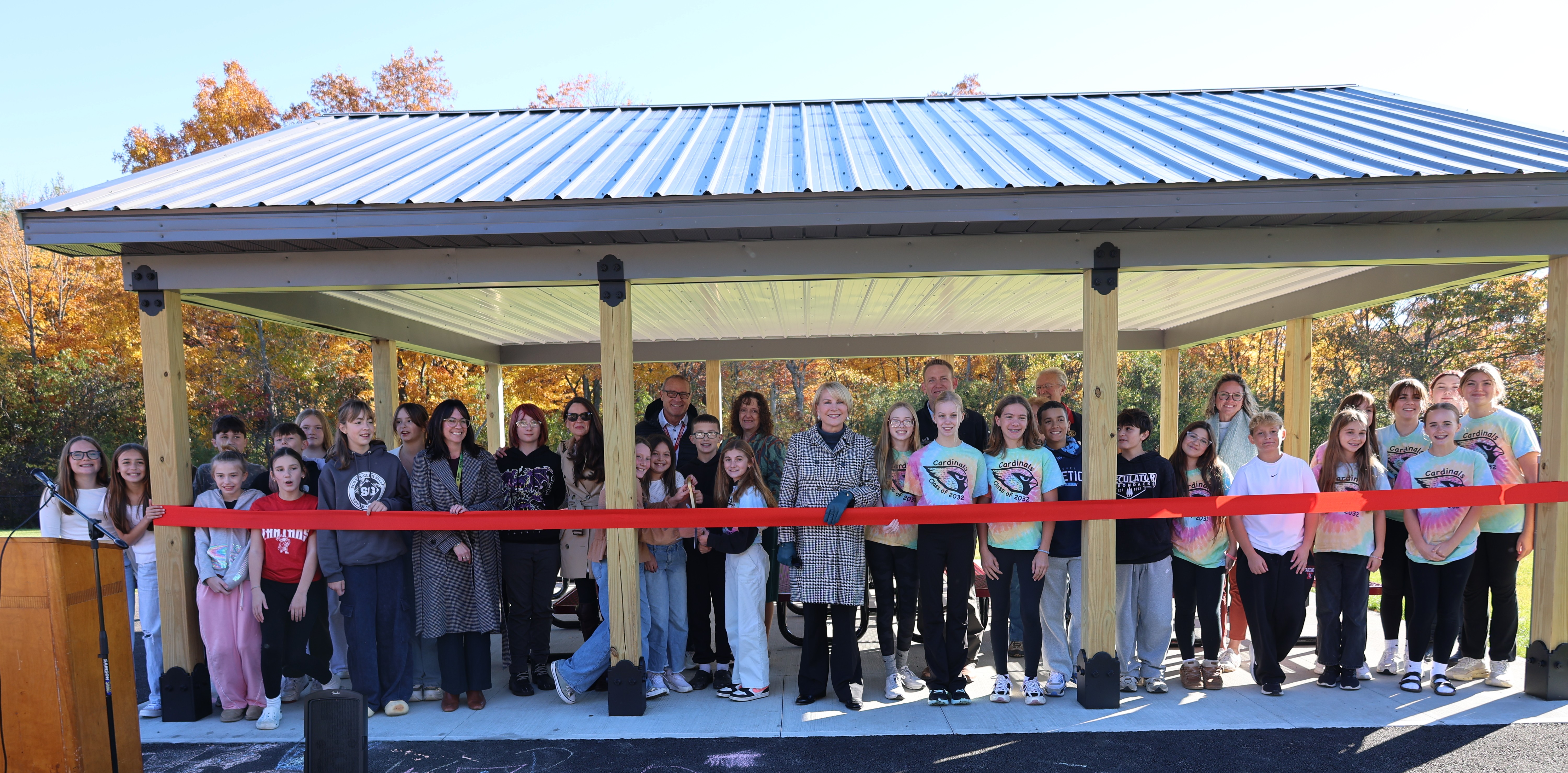 A group of students, teachers, and community members gather under a pavilion to celebrate with a red ribbon-cutting ceremony.