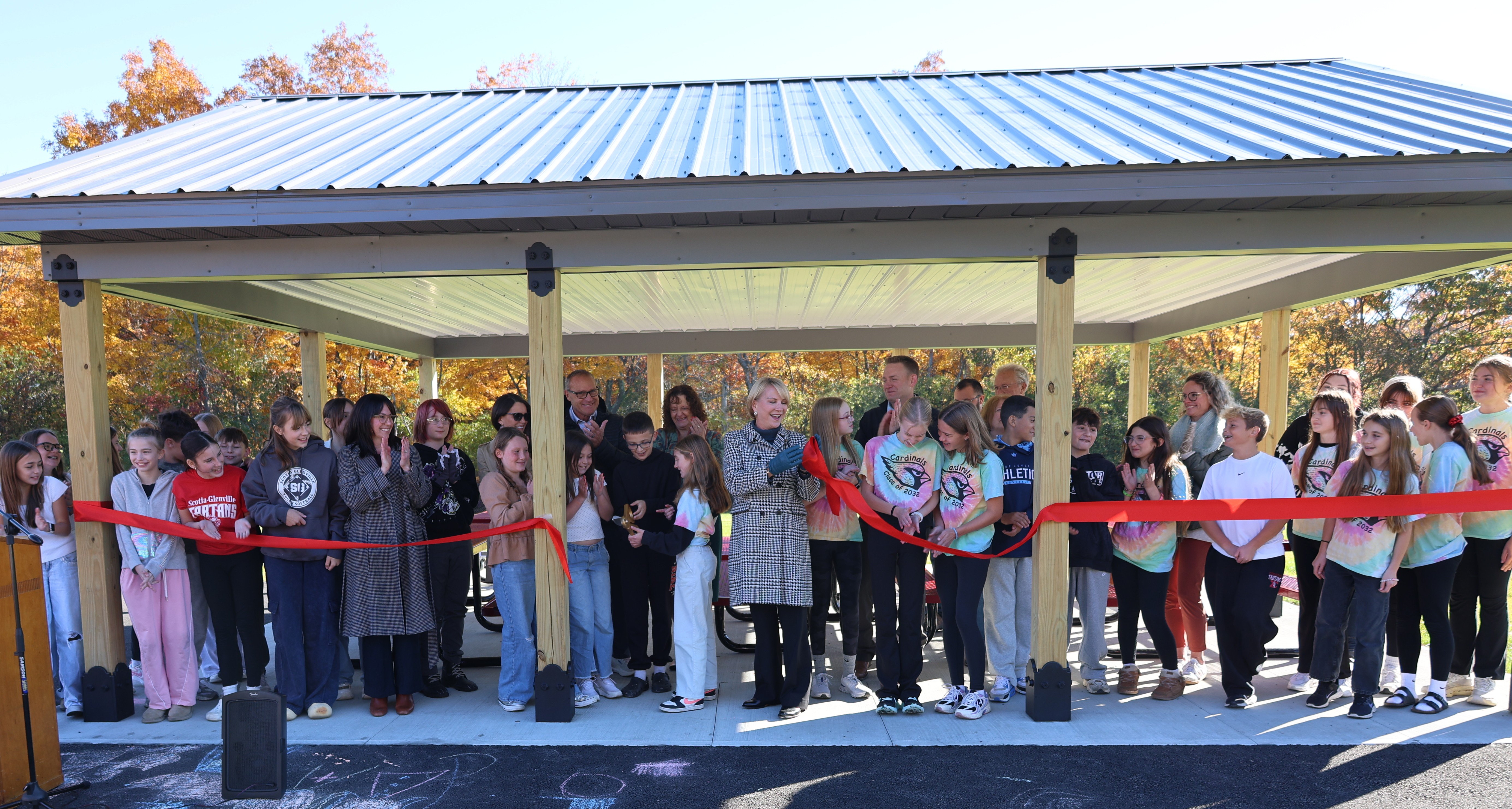 A group of students, teachers, and community members gather under a pavilion to celebrate a ribbon-cutting ceremony outdoors.