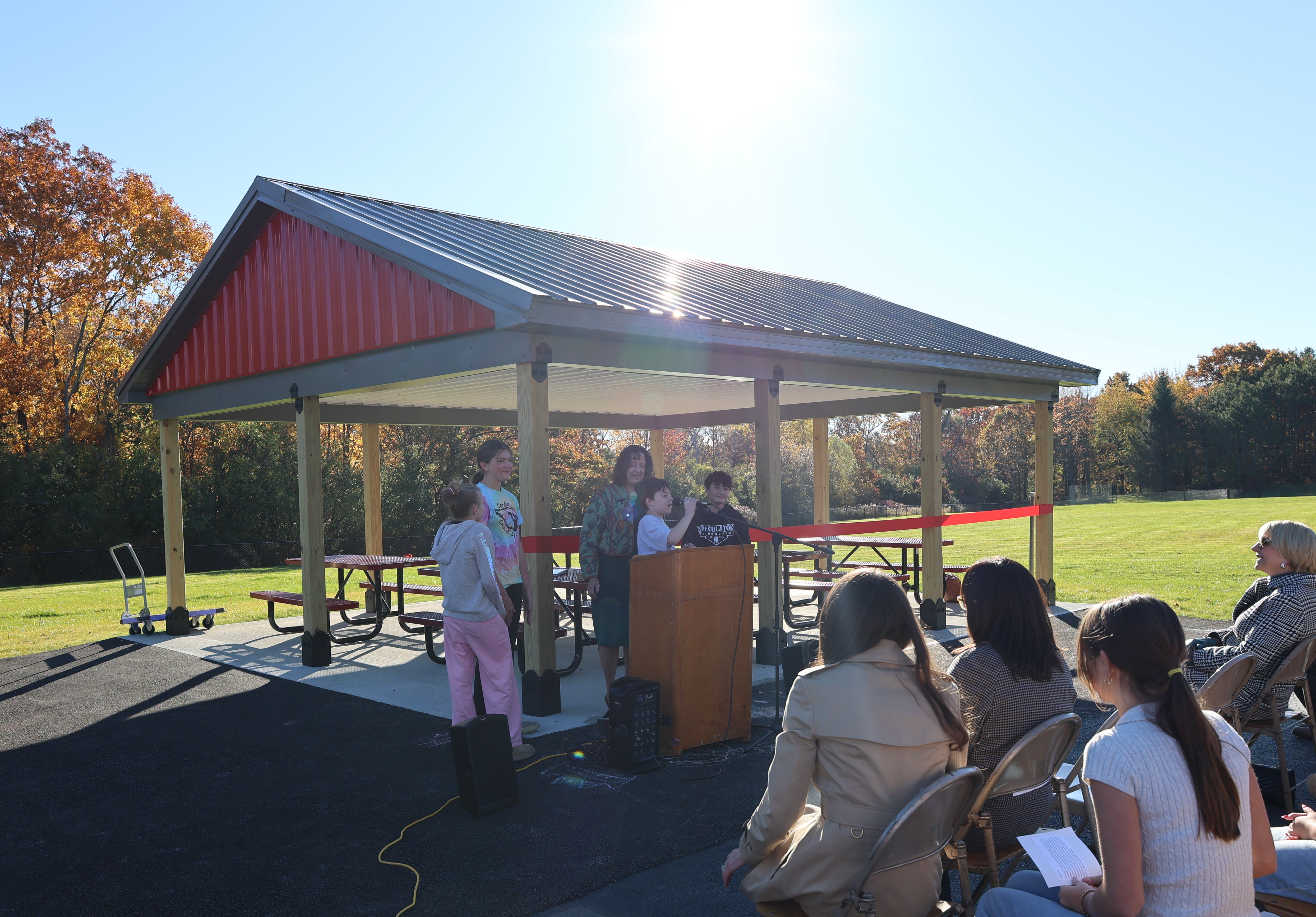 A group of people gathers under a pavilion for an outdoor presentation on a sunny day.