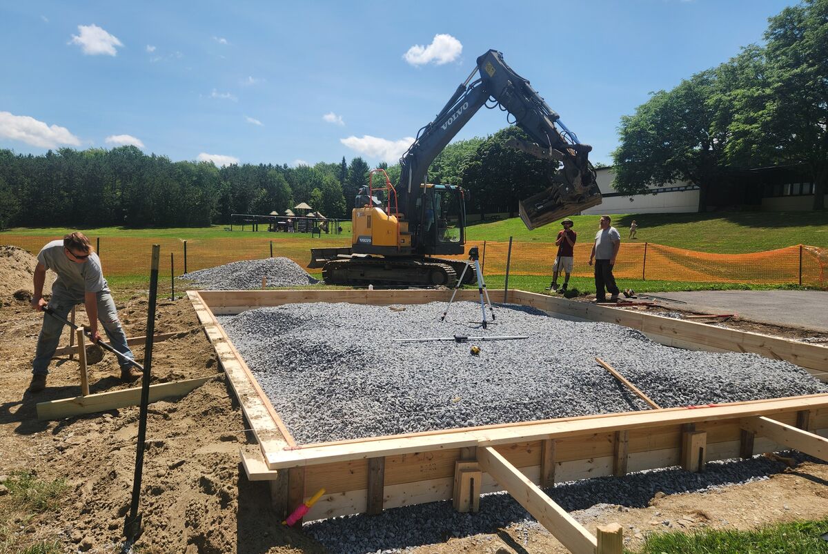 A worker shovels dirt near a wooden foundation filled with gravel, while an excavator operates in the background.