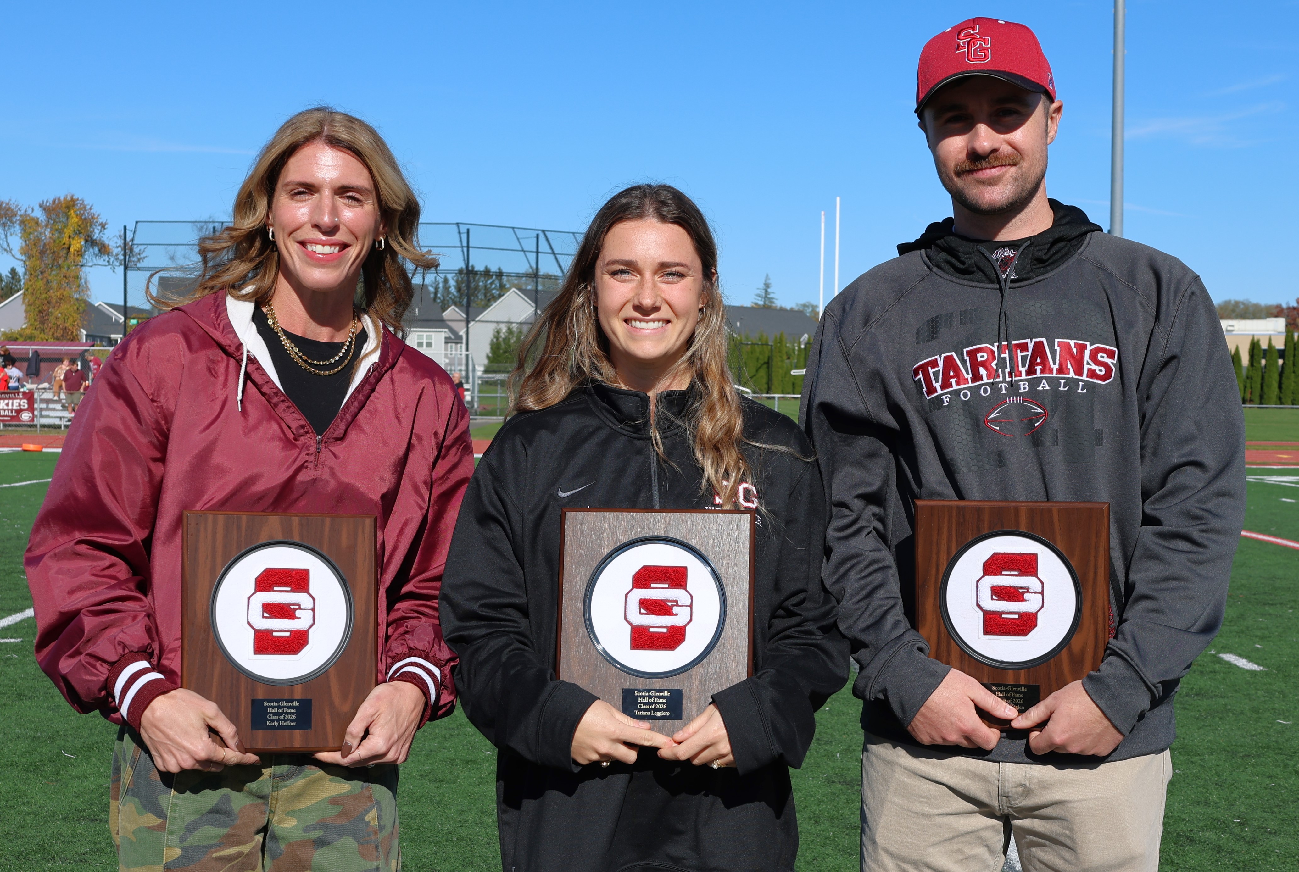 Three people proudly hold plaques featuring the red letter G logo at an outdoor sports field on a sunny day.