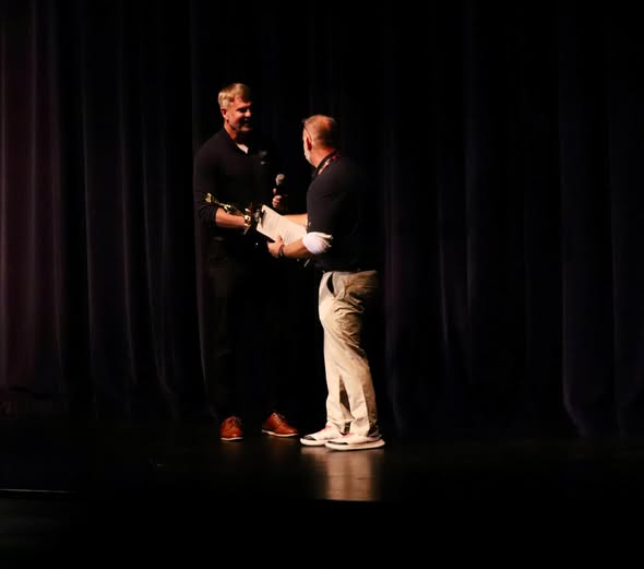 A presenter on stage hands an award to a recipient in a dimly lit auditorium.