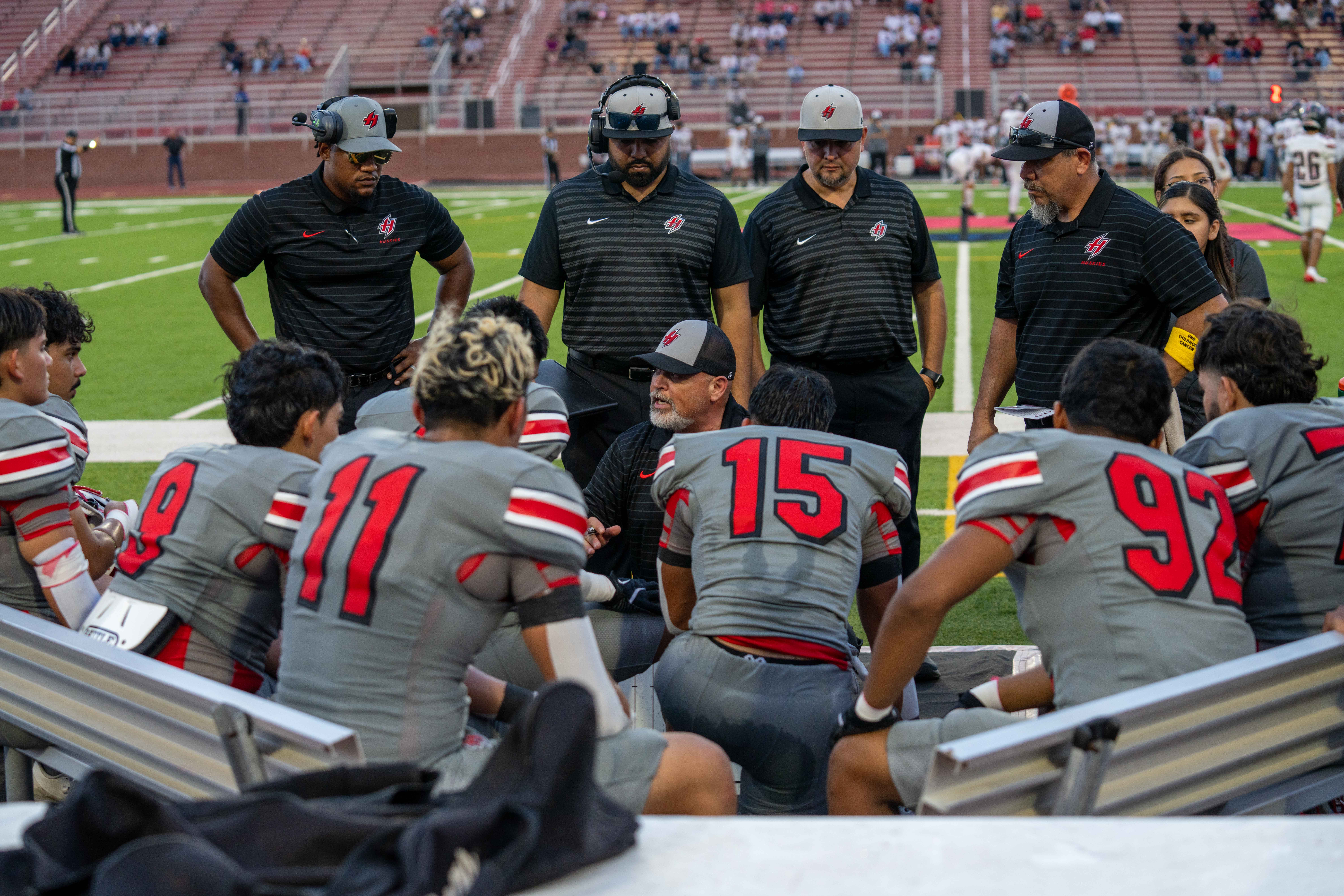 A football coach crouches to talk to his team, which sits in a huddle on the sidelines.