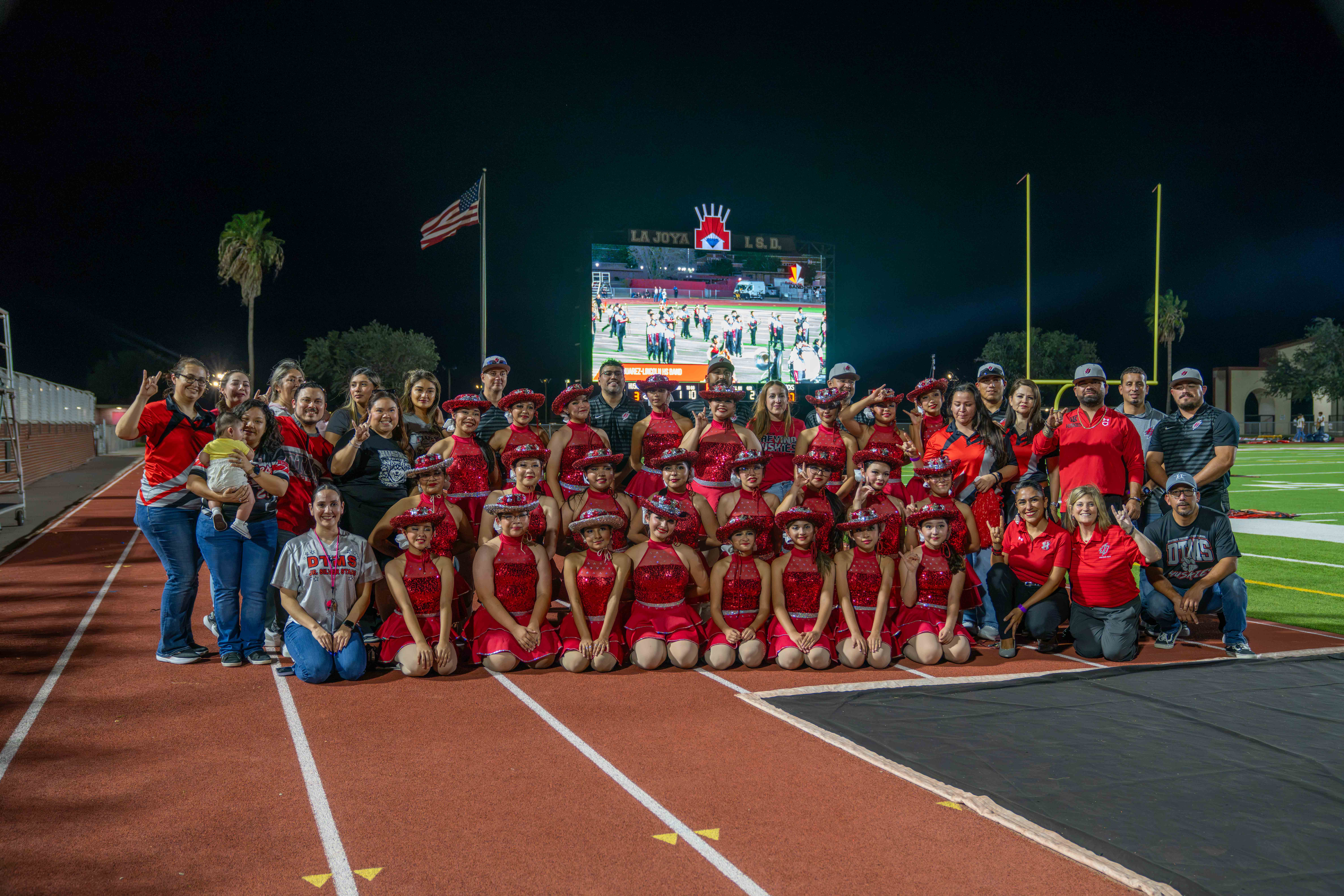 A large group of youth dancers in matching red sequin outfits and hats pose on a running track at night, accompanied by family and staff.
