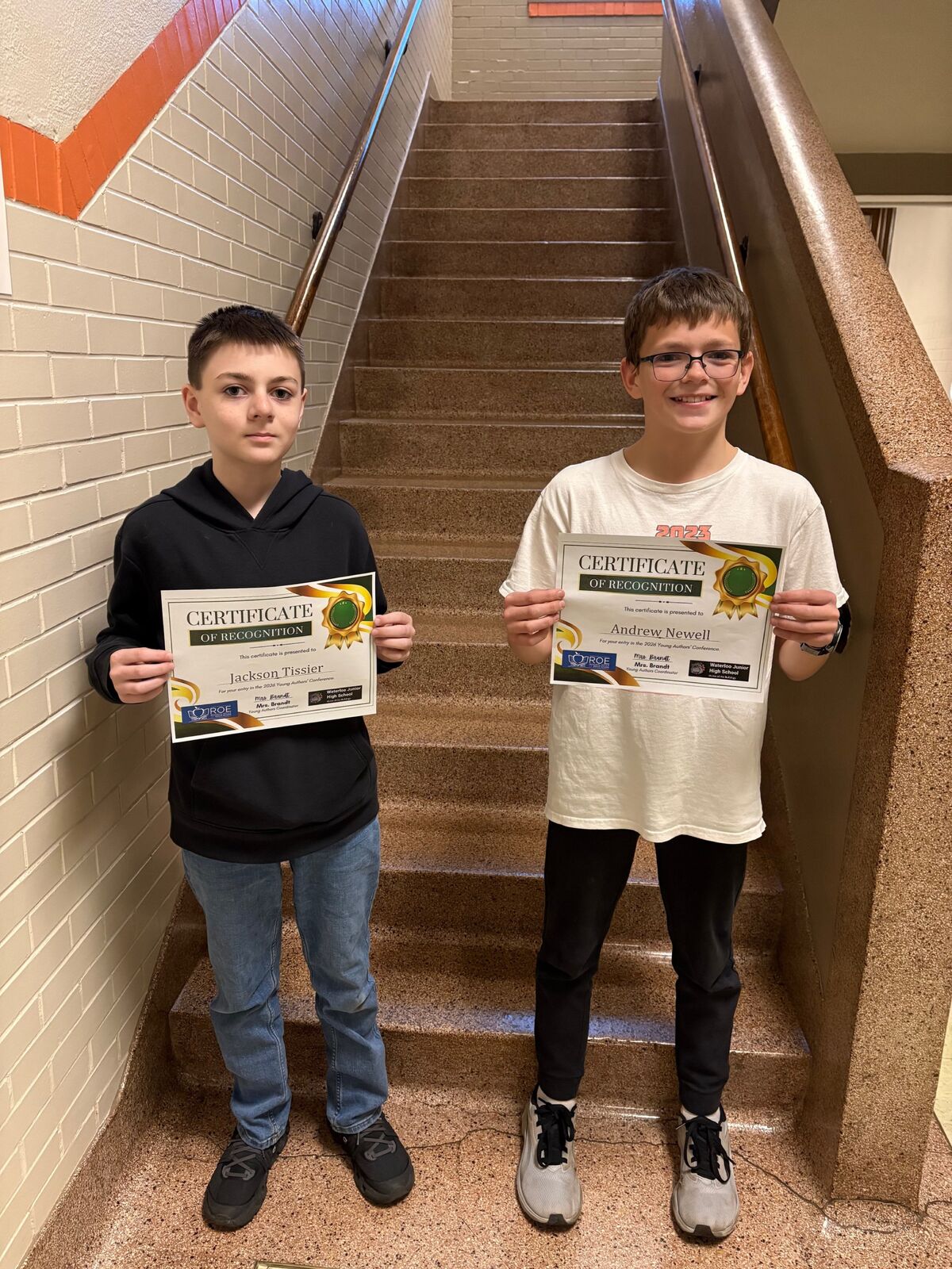 Two boys stand on stairs holding certificates of recognition for the 2026 Young Authors' Conference.