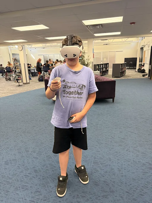 A child wearing a VR headset and holding controllers stands in a library or media center.