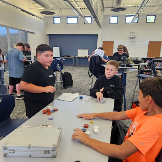 Students gather around a table in a bright classroom to play a game with colorful tokens and a metal case.