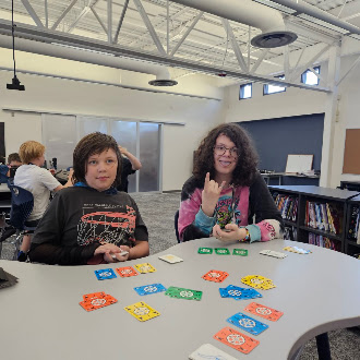 Two students sit at a curved table in a library, playing a card game with colorful cards.