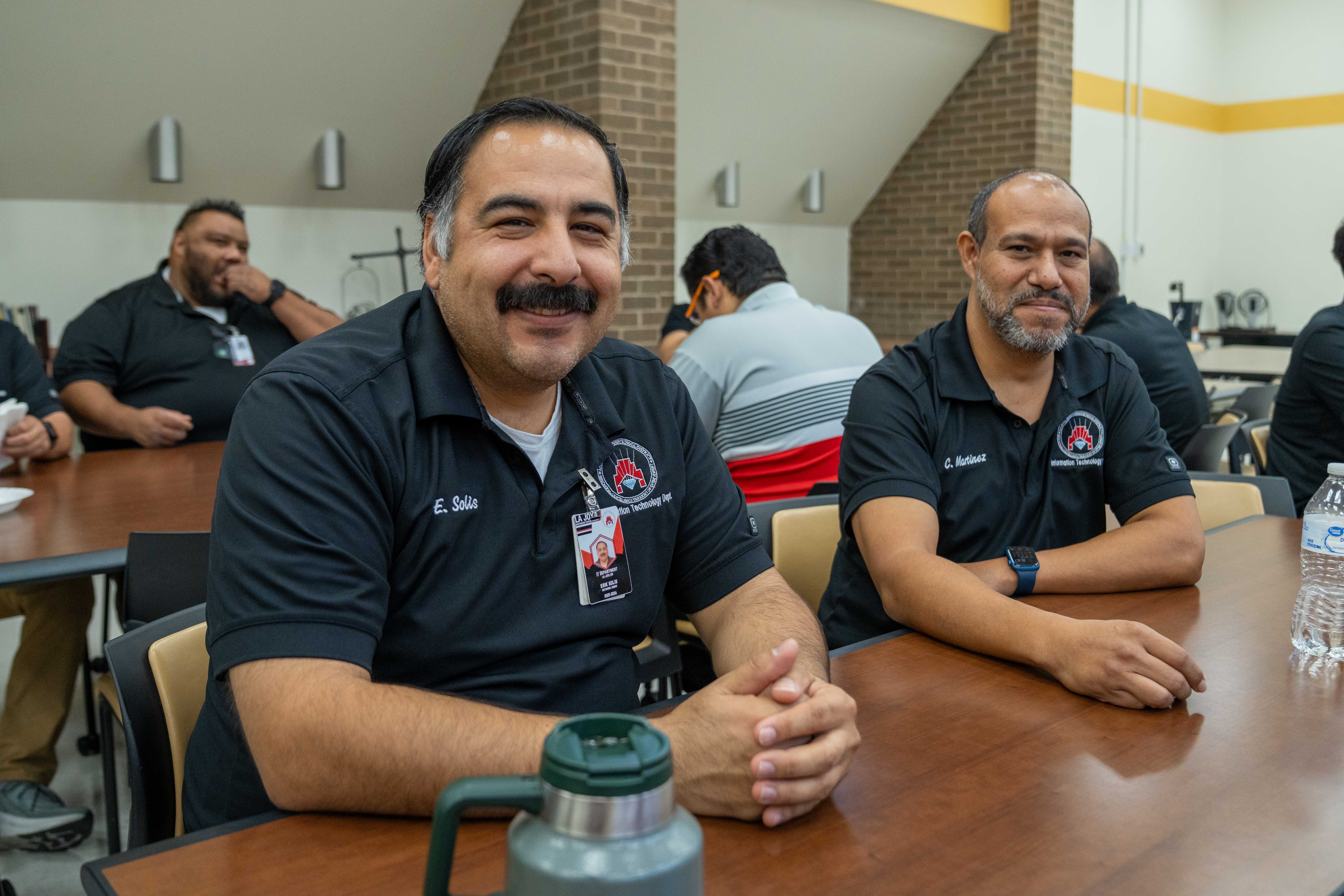 Two men in matching black polo shirts sit at a wooden table, smiling at the camera indoors.
