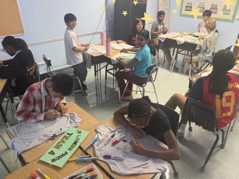 Students sit at desks in a classroom, focused on decorating gray t-shirts with markers.
