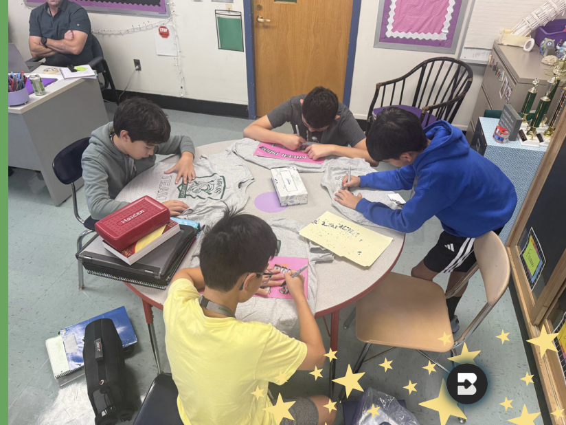 Four students sit around a round table in a classroom, focused on signing gray t-shirts.