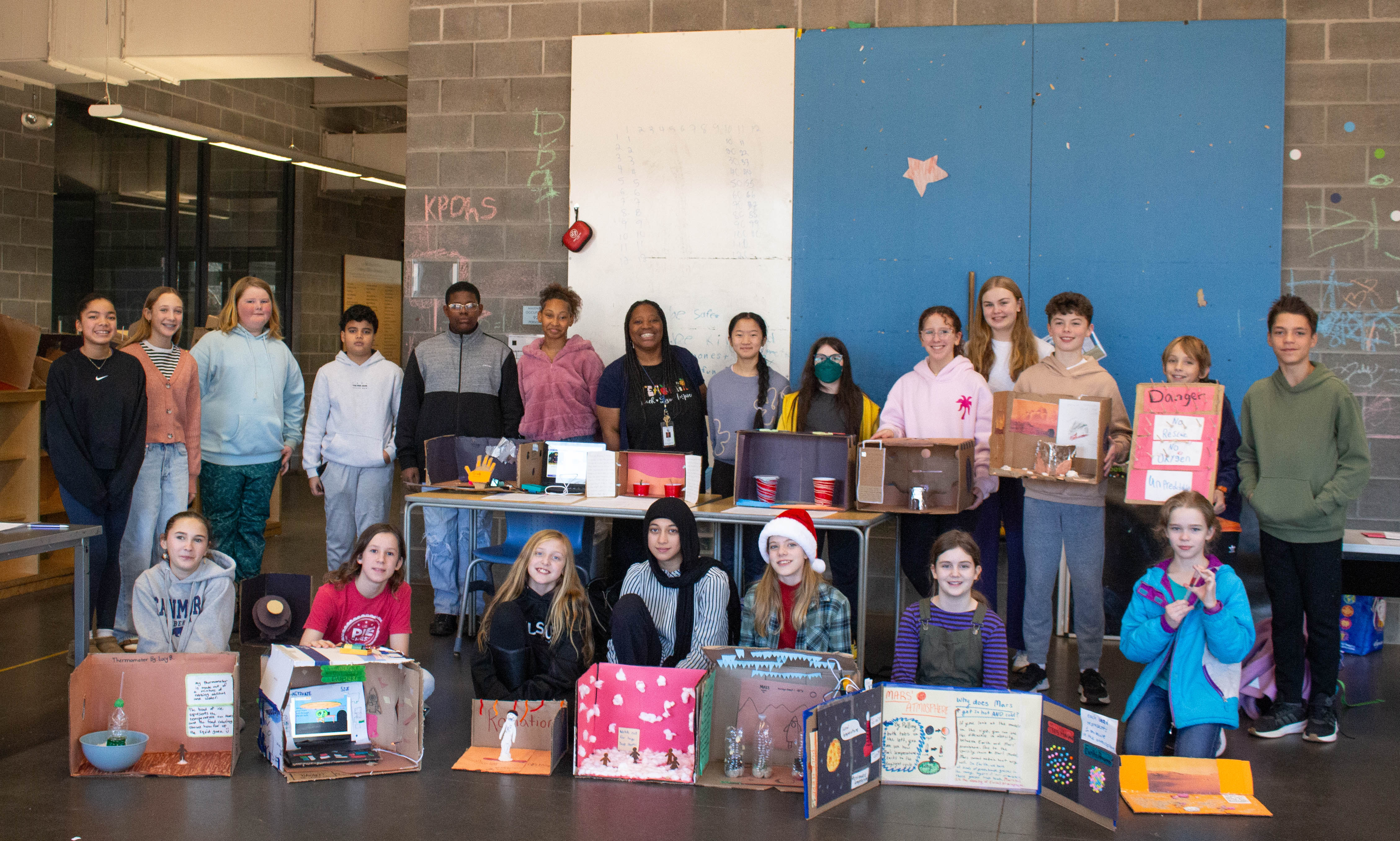 A diverse group of students stands behind their science projects, which are displayed on the floor and tables.