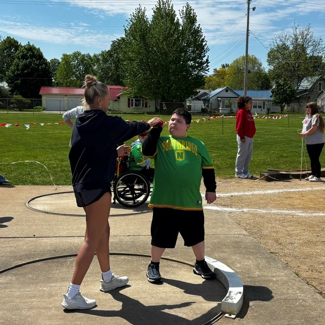 A young woman assists a boy in a green shirt with a shot put during an outdoor track and field event.