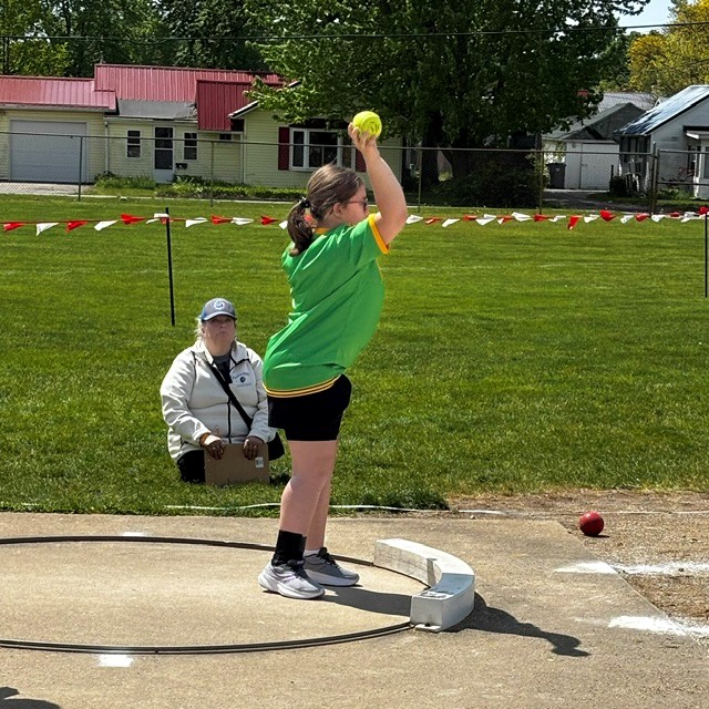 A young athlete in a green shirt prepares to throw a yellow ball in a shot put event.