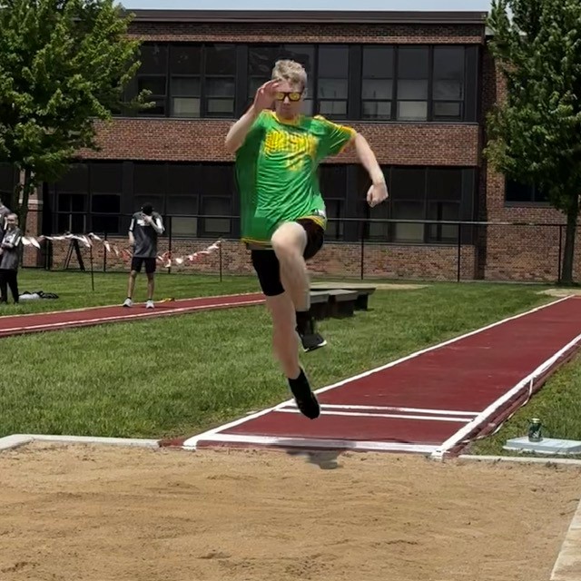A young athlete in a green shirt leaps during a long jump competition on a sunny day.