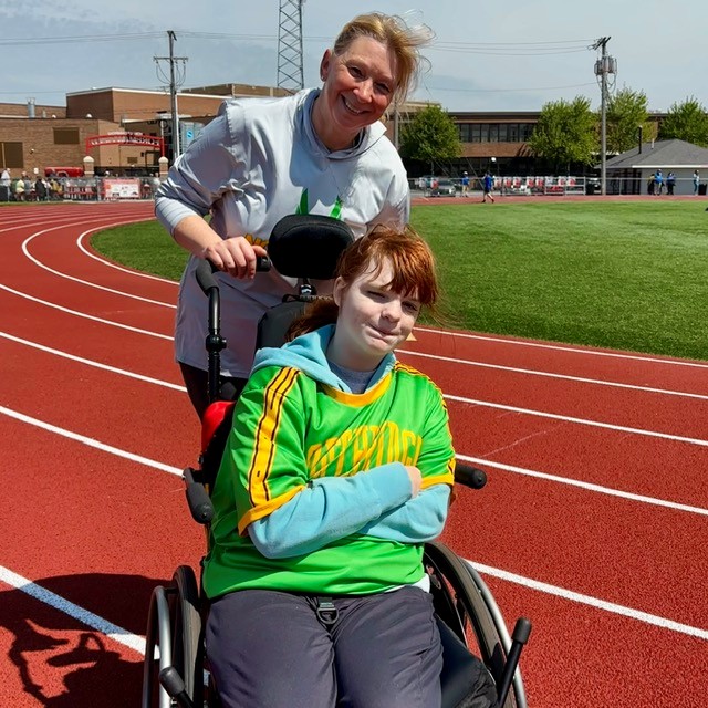 A woman pushes a young person in a wheelchair on a red running track.