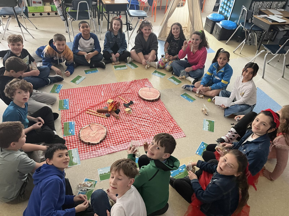 Children sit in a circle on the floor around a red and white checkered blanket with a pretend campfire and logs.