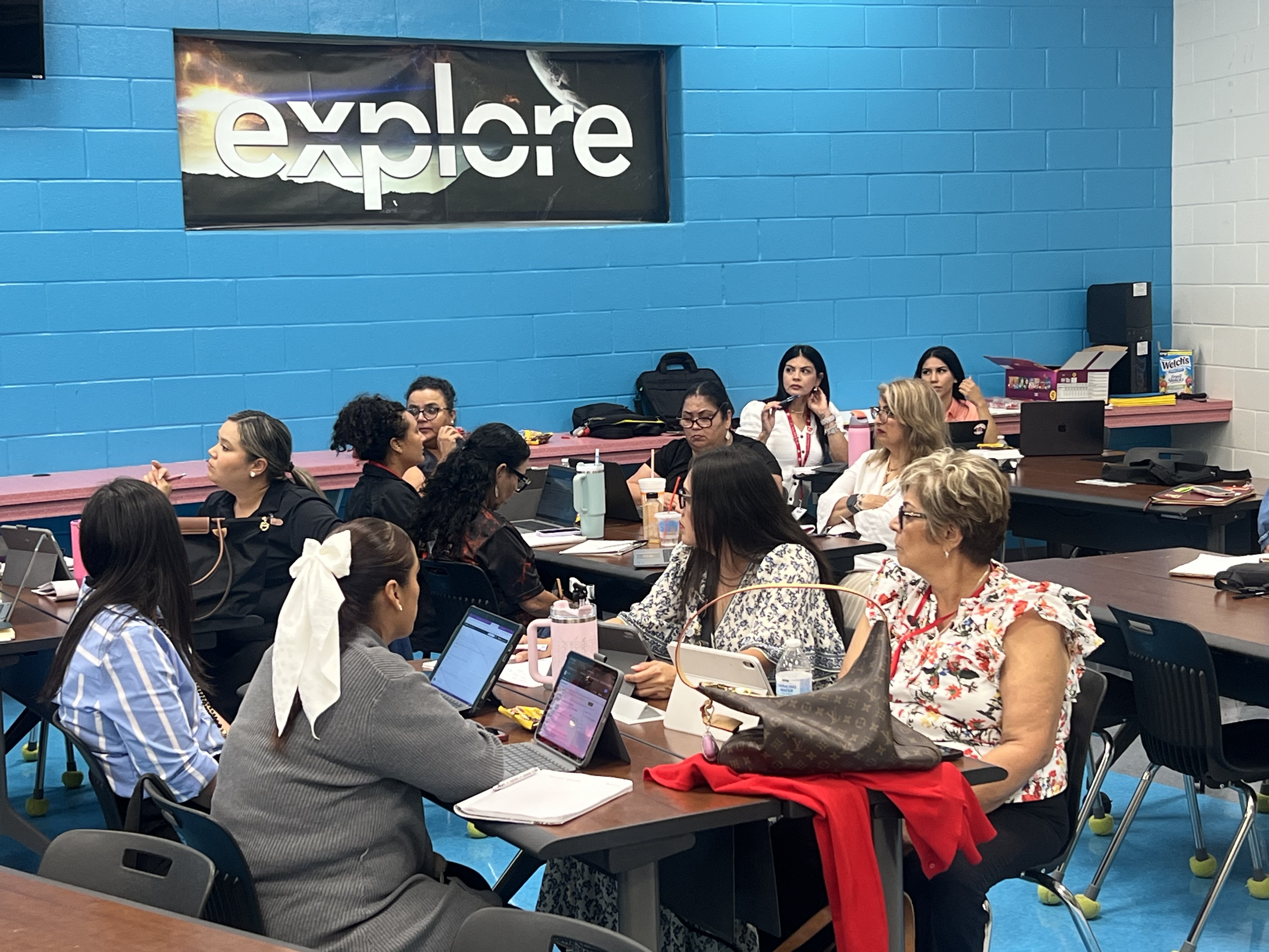 A group of women are seated at tables in a classroom, engaged in a presentation or training session.
