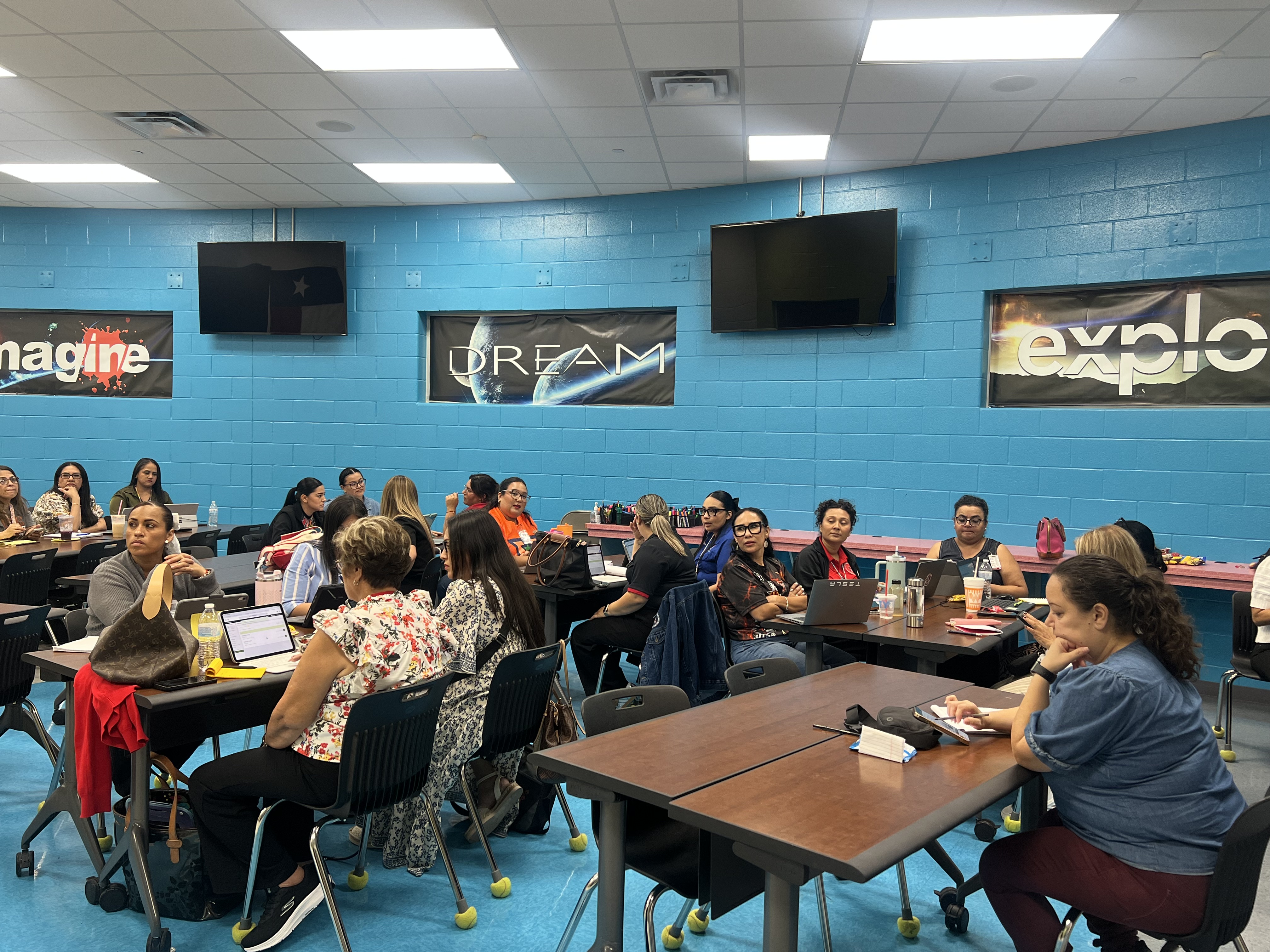 A group of adults are seated at tables in a classroom, engaged in a presentation or training session.