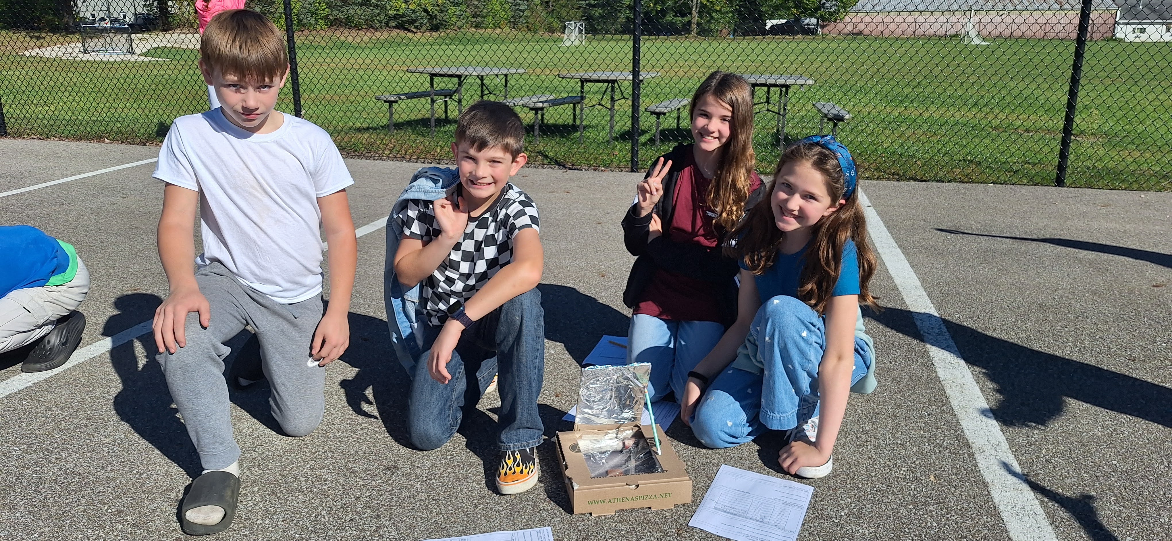 Four children pose outdoors on an asphalt playground next to a science project made from a pizza box.