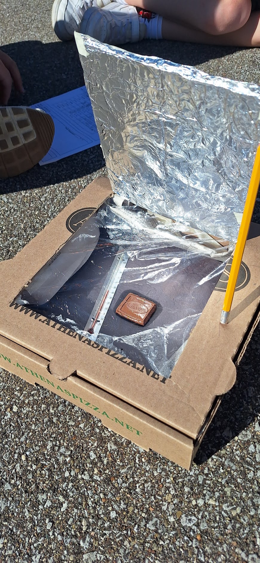 A DIY solar oven made from a pizza box sits on asphalt, containing a thermometer and a piece of chocolate.