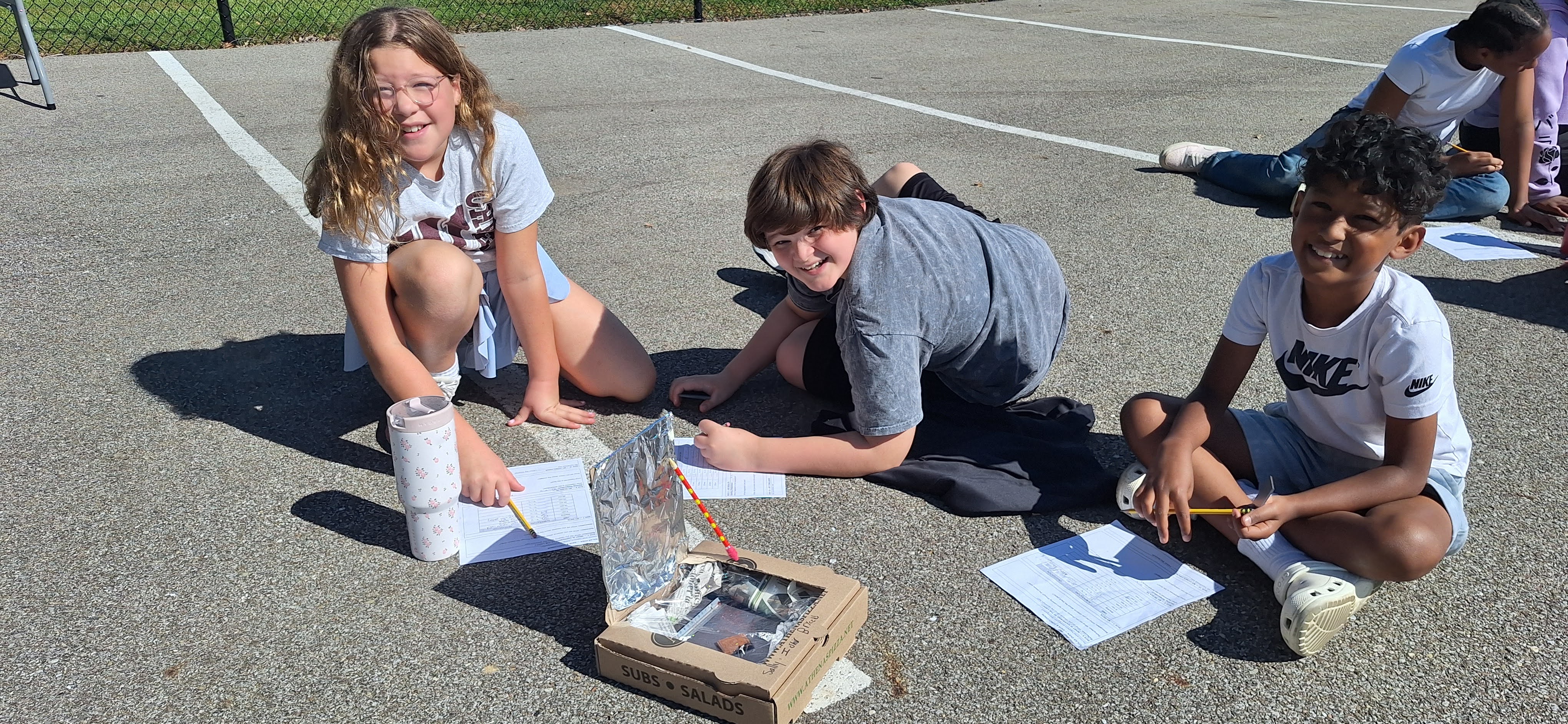 Three children sit on an outdoor asphalt parking lot, smiling and working on a science project involving a homemade solar oven built from a pizza box.