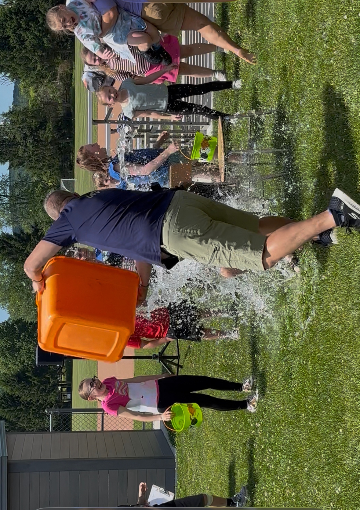 A man pours water from an orange container onto people sitting on chairs, creating a splash.