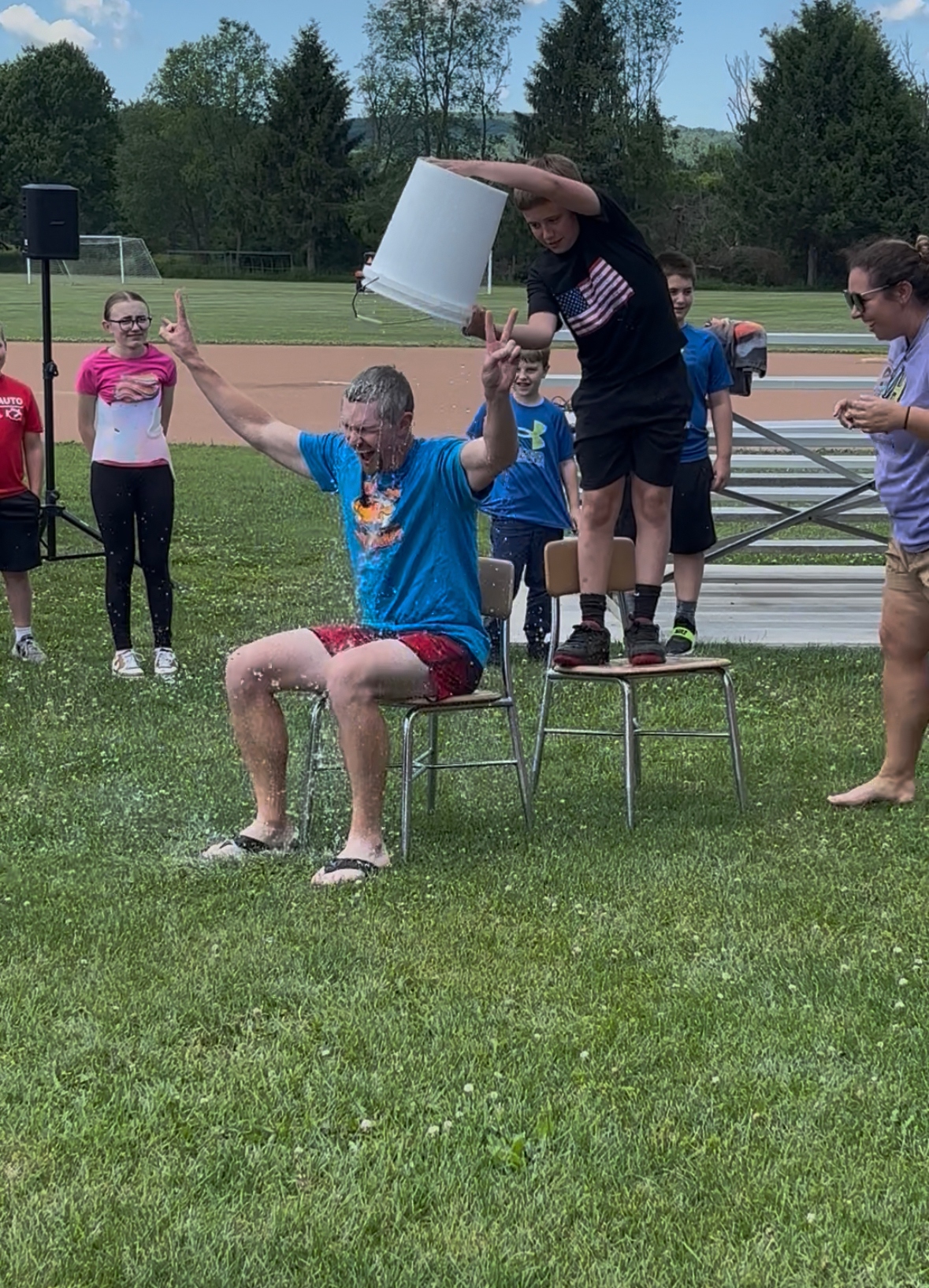 A man sits on a chair outdoors as a bucket of water is poured over his head by a boy.