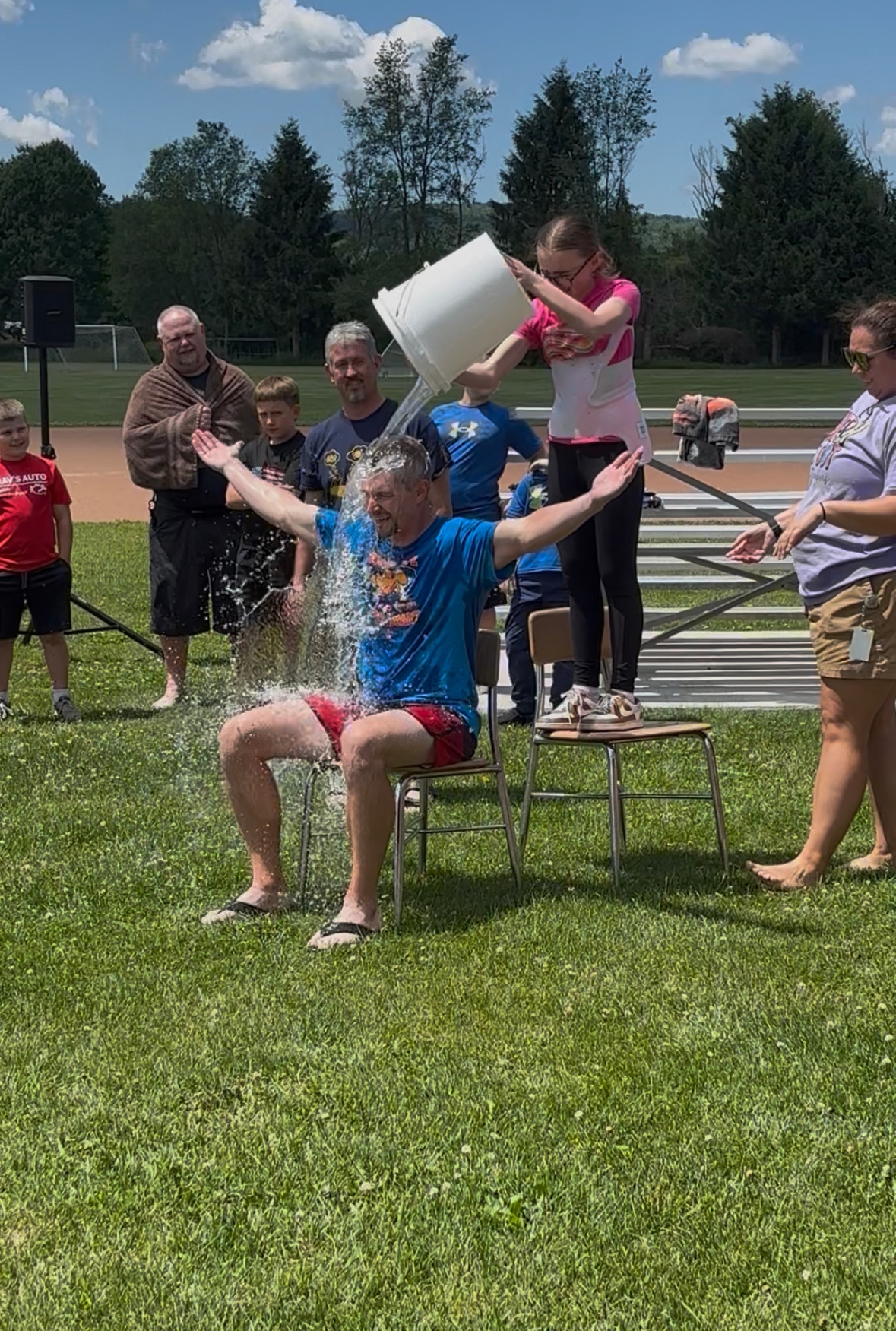 A man sits on a chair outdoors as a bucket of water is poured over his head by a young girl.