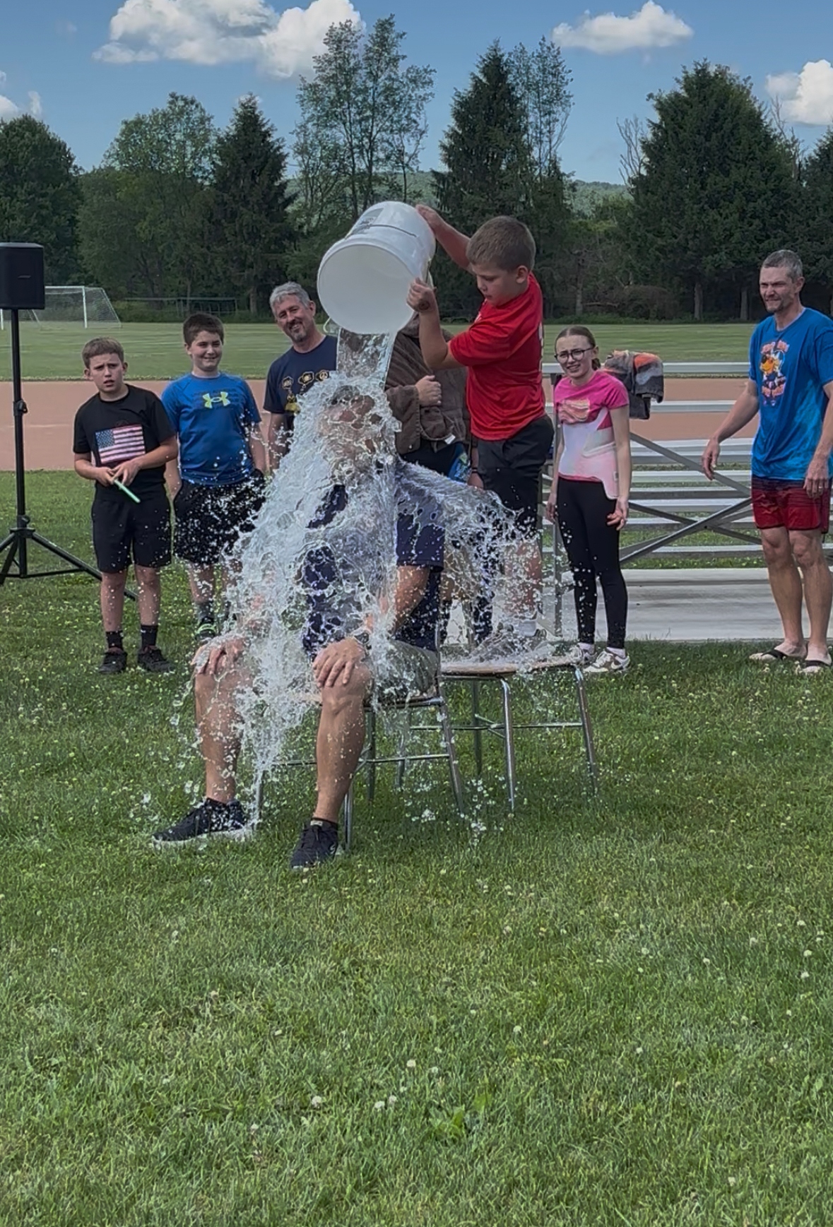 A boy pours a bucket of water over a man sitting on a chair outdoors.