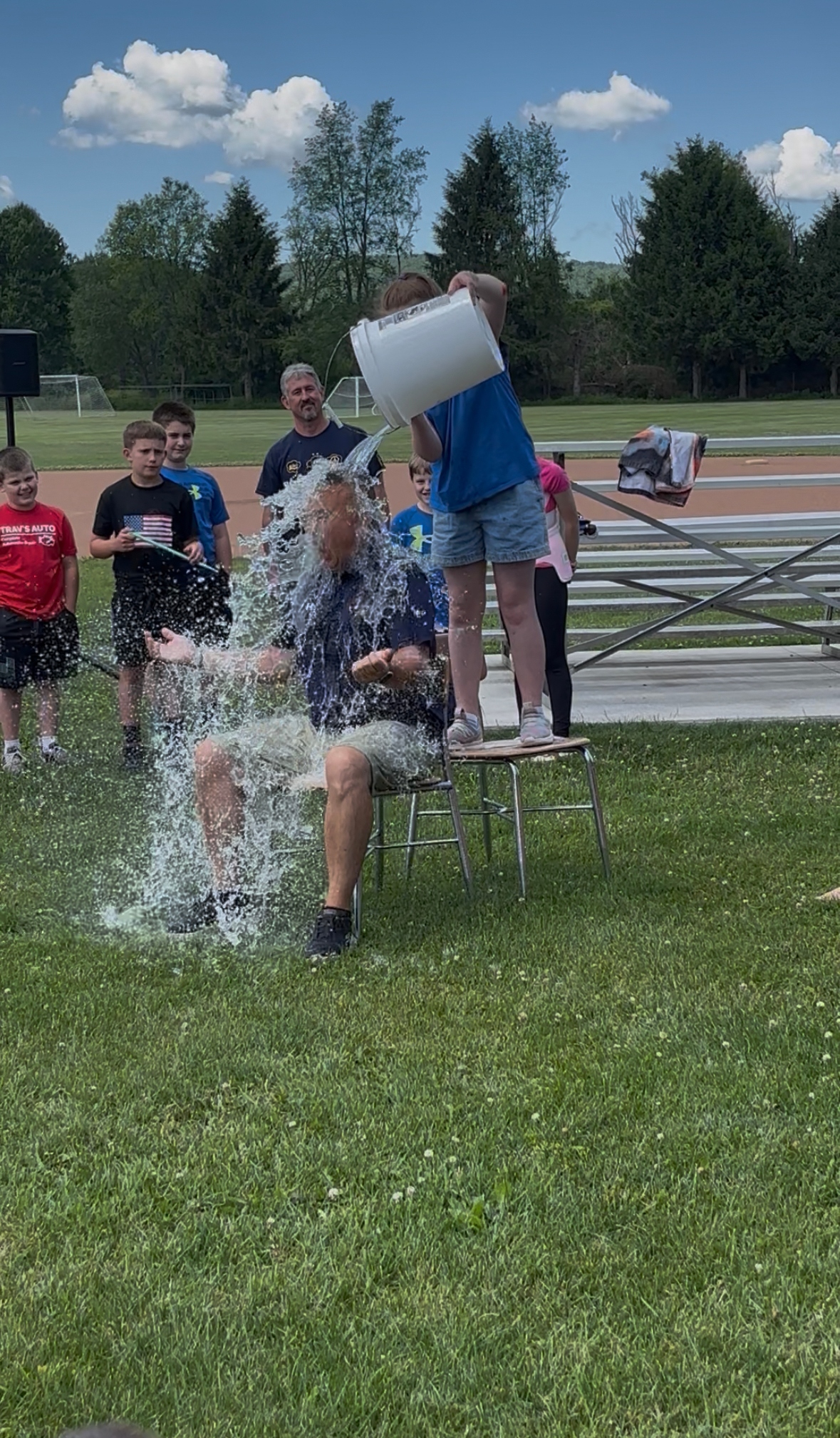 A person is doused with a bucket of water while sitting on a chair outdoors, surrounded by onlookers.