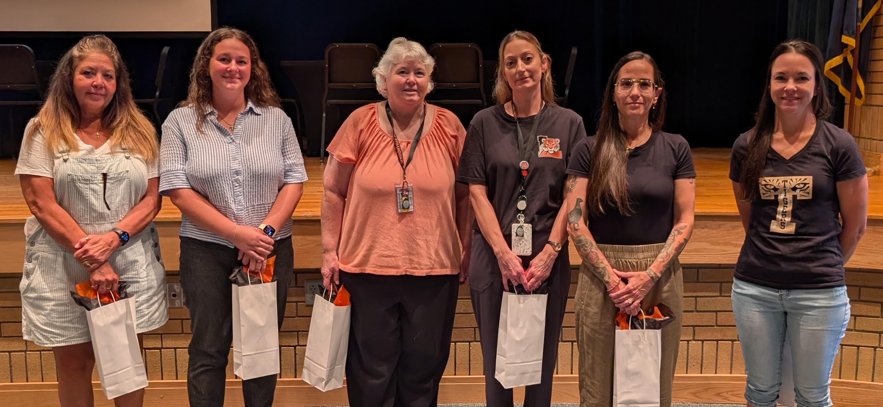 Six women stand on a stage holding white gift bags, posing for a group photo indoors.