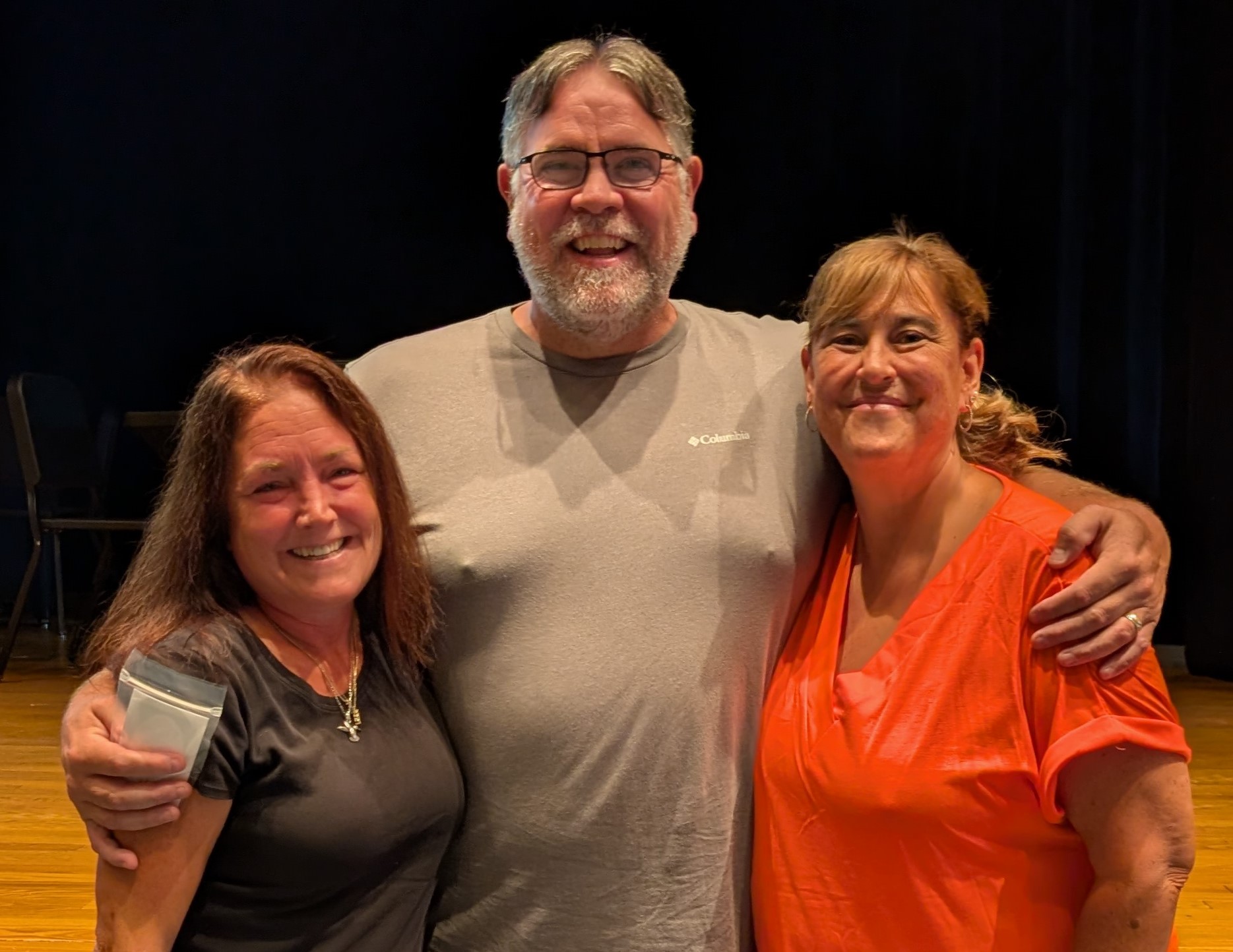 Three smiling adults stand closely together, posing for a photo indoors on a wooden stage.
